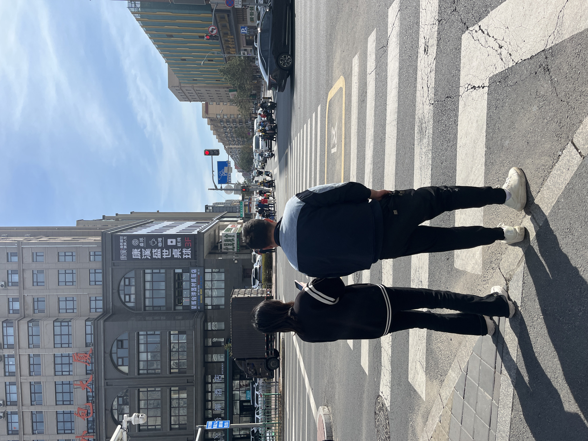The image depicts a street scene in Changchun City, China. Two individuals, a man and a woman, are crossing a zebra crossing. The man is wearing a dark blue jacket with a light blue panel, black pants, and white sneakers. The woman is wearing a black jacket with white stripes on the sleeves and black pants with black and white sneakers. They are facing away from the viewer.
In the background, there is a multi-story building with several windows and advertisements in Chinese characters. Above the road, a traffic light is visible, showing a red signal. Cars and motorcycles are lined up waiting behind the crosswalk. The sky is partly cloudy. The zebra crossing is cracked and shows signs of wear.