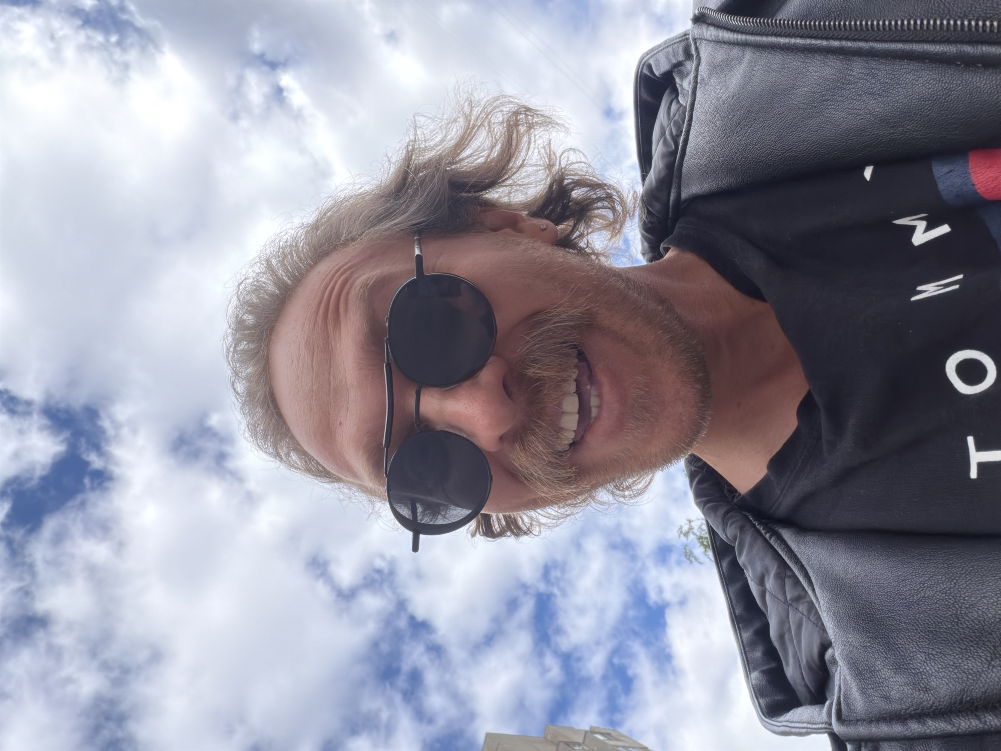 A man wearing sunglasses, a black t-shirt, and a black leather jacket, is looking down at the camera from a low angle. He has blond hair and a mustache. The sky with white clouds is in the background. A building is visible in the lower left corner. The photograph was taken in Saint Petersburg, Russia.