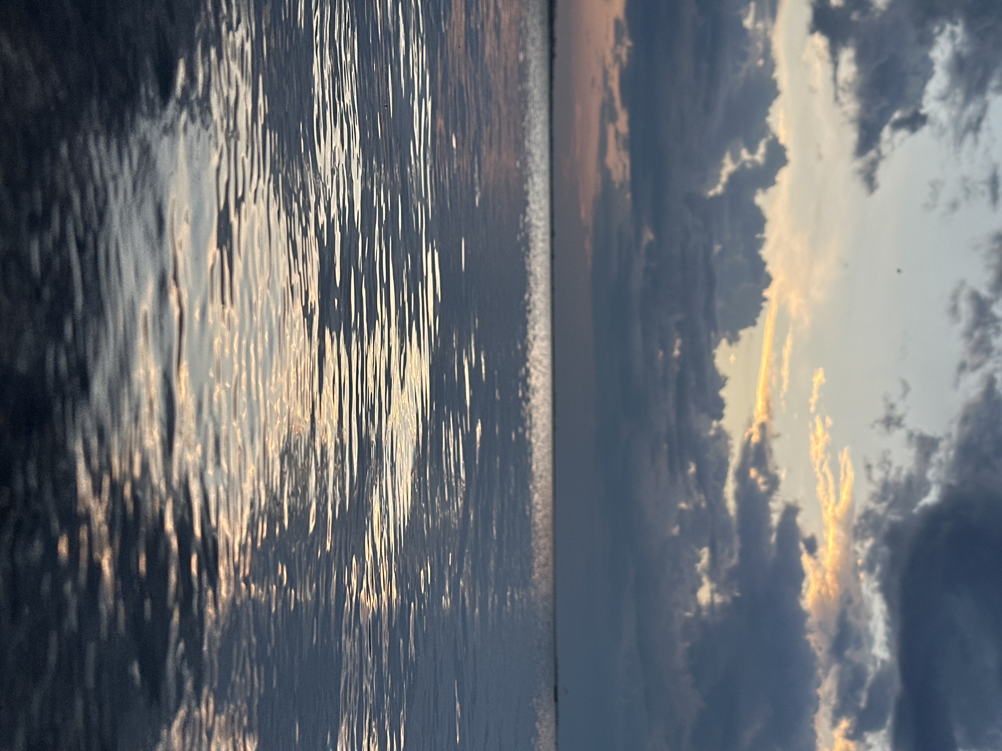 The image depicts a body of water under a cloudy sky, likely at dusk or dawn. The sky is filled with dark, heavy clouds interspersed with streaks of yellow and orange light, suggesting the sun is low on the horizon. The water's surface is rippled, reflecting the colors of the sky and appearing somewhat metallic. A distant, hazy horizon line separates the water from the sky. The location is Kokshetau, Kazakhstan.