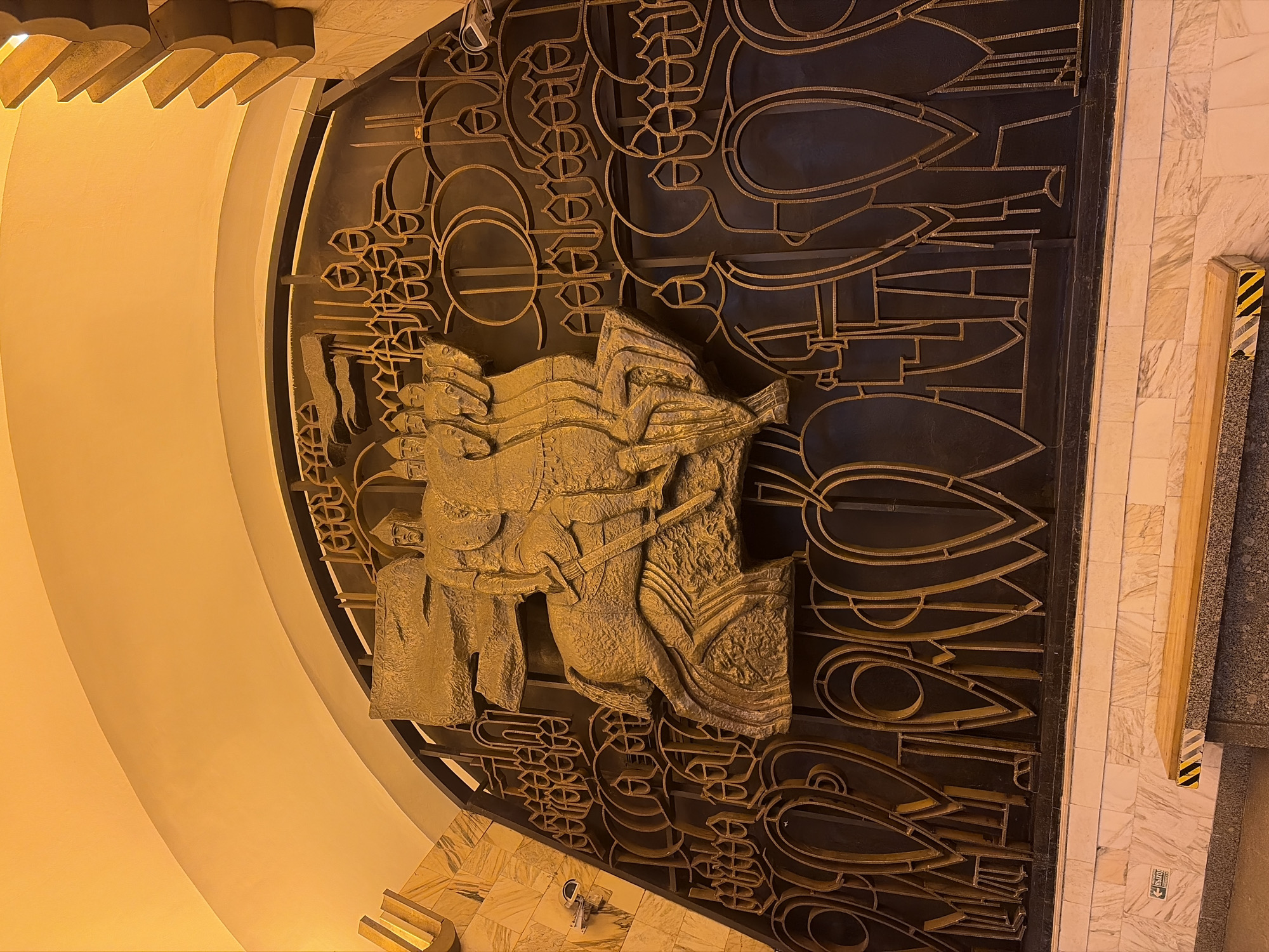 The image shows a large metal relief sculpture mounted on a wall or gate. The sculpture is primarily gold/bronze in color, with intricate details. In the center is a depiction of figures. These figures appear to be from the medieval or classical era. The wall or gate is decorated with abstract geometric and organic shapes. The background suggests an interior space with beige/light brown walls and floors. The location is Saint Petersburg, Russia. The artwork is likely located in the Avtovo metro station.