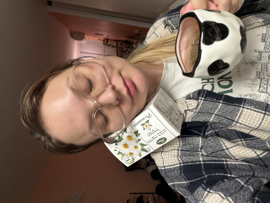 A person with glasses is lying down with closed eyes, holding a cow-patterned mug of tea. A box of "Romashka" chamomile tea rests near their head. The person is wearing a white t-shirt and a plaid shirt. The background shows a glimpse of a room with a hallway.