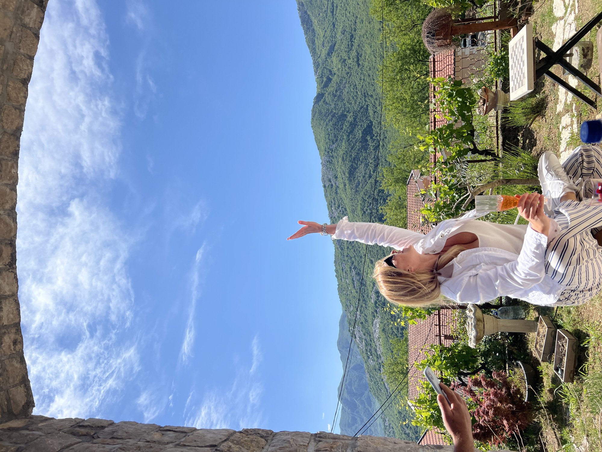 A woman is seated outdoors on a sunny day in Sotonići, Montenegro, holding a drink in one hand and extending the other towards a backdrop of mountains and sky. She is wearing a white shirt, sunglasses, and striped pants. The scene is framed by a stone structure in the foreground. A hand is holding a phone. Additional objects include a chess board, a table, plants, and various decorations, typical of a garden or patio setting.