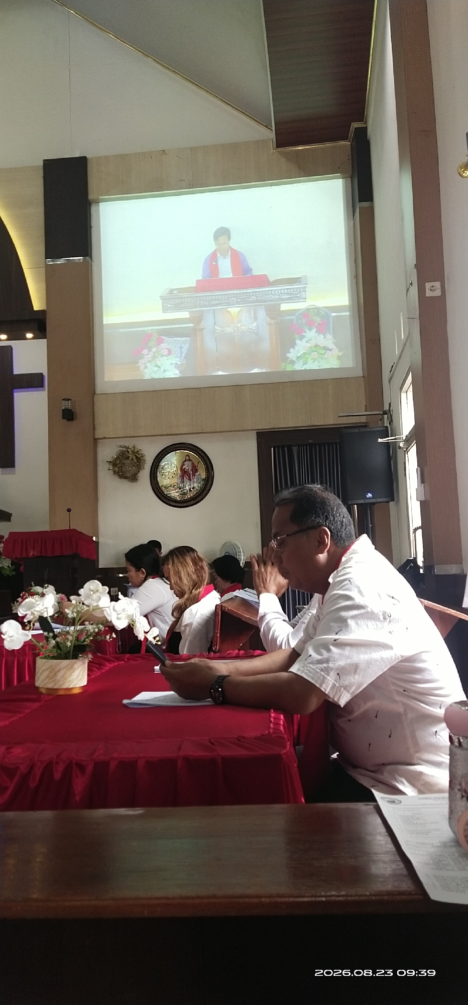 The image depicts an indoor scene within what appears to be a church or a religious hall, possibly located in the city of Jaar, Indonesia, as indicated by the timestamp. A projector screen at the front displays a person in religious attire standing behind a lectern. In the foreground, several individuals are seated at a long table draped with a red tablecloth. A man in the right foreground, wearing glasses and a white shirt with a subtle pattern, is looking down with his hands clasped in front of him, holding a mobile phone. To his left, other individuals, predominantly women, are also seated, some with red stoles around their necks, suggesting a uniform or ceremonial dress. The table is adorned with floral arrangements. The overall ambiance suggests a formal gathering or service.