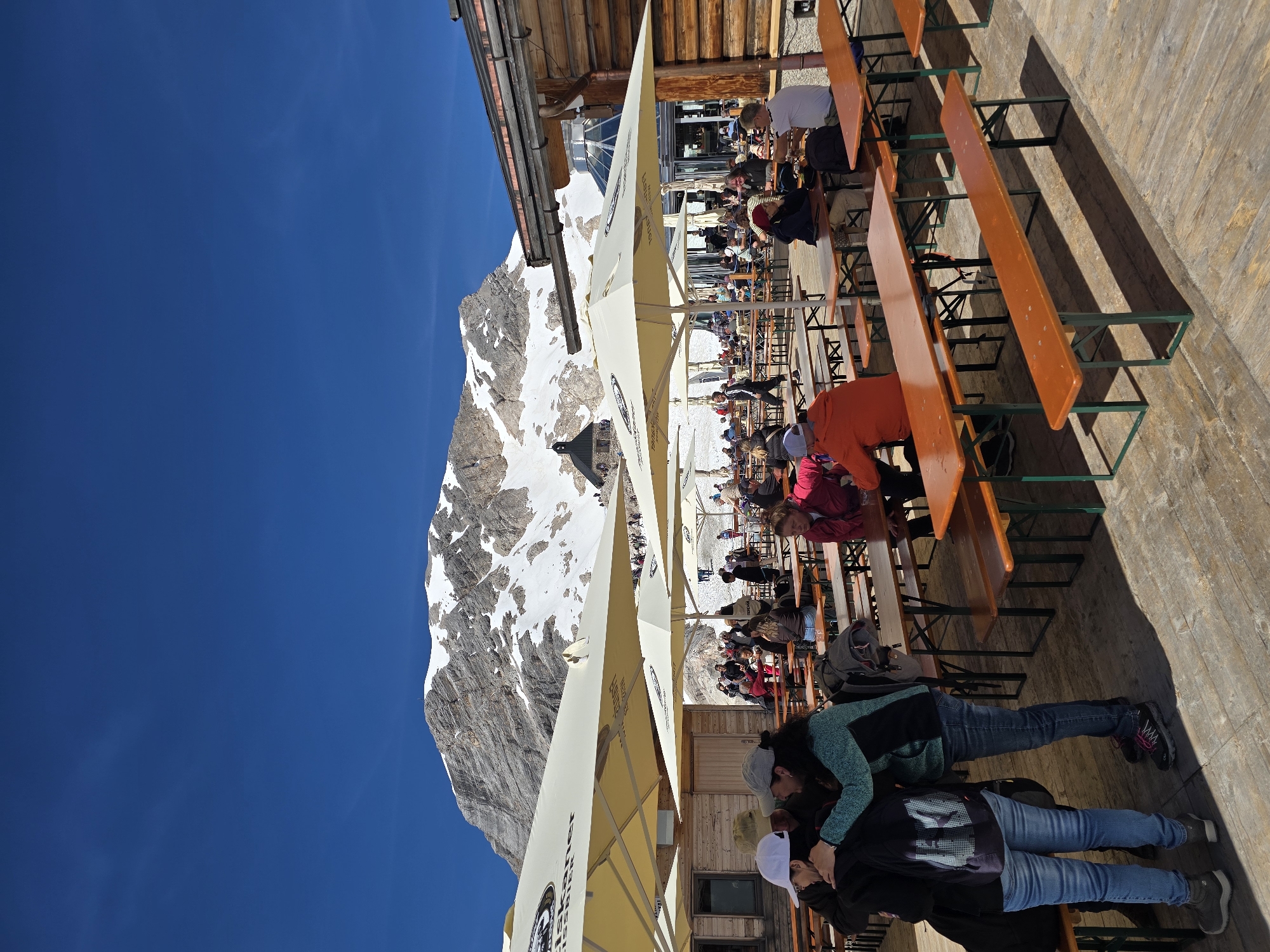 The image depicts an outdoor seating area with numerous picnic tables and benches, covered by large white umbrellas, set against the backdrop of a snow-capped mountain under a clear blue sky. The location is identified as the Zugspitze mountain, the highest peak in Germany, near Garmisch-Partenkirchen. People are seated at the tables, some eating and drinking, while others are standing and conversing. A wooden building, likely a restaurant or cafe, is visible to the right, with a glass dome structure and a church-like building on the mountain in the distance. The ground is a wooden deck. The overall atmosphere is one of a busy, popular tourist destination.