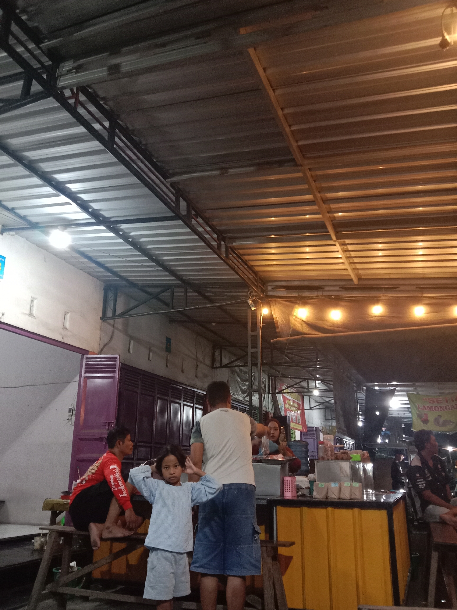The image depicts an outdoor food stall with a corrugated metal roof supported by metal beams. The lighting appears to be artificial, with several yellow lights illuminating the scene from above. In the foreground, a young child in a light blue shirt and shorts stands with their hands near their head, looking towards the camera. To the left of the child, an adult in a red shirt and black pants is seated on a wooden bench. Behind the child, another adult in a white shirt and blue shorts stands. Further back, a woman is visible behind a yellow counter, appearing to be serving customers. There are several other individuals in the background, some seated at tables and others standing. Bags of snacks or goods are displayed on the counter. The environment suggests a casual, possibly nighttime, setting for a food establishment in Cangkring, Indonesia.