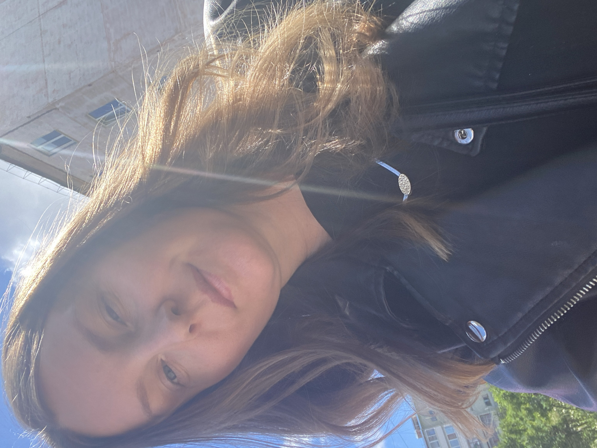 A woman with light brown hair, wearing a black leather jacket, is taking a selfie outdoors. The backdrop includes buildings and trees, with a bright sky suggesting a sunny day in Noviy, Russia.