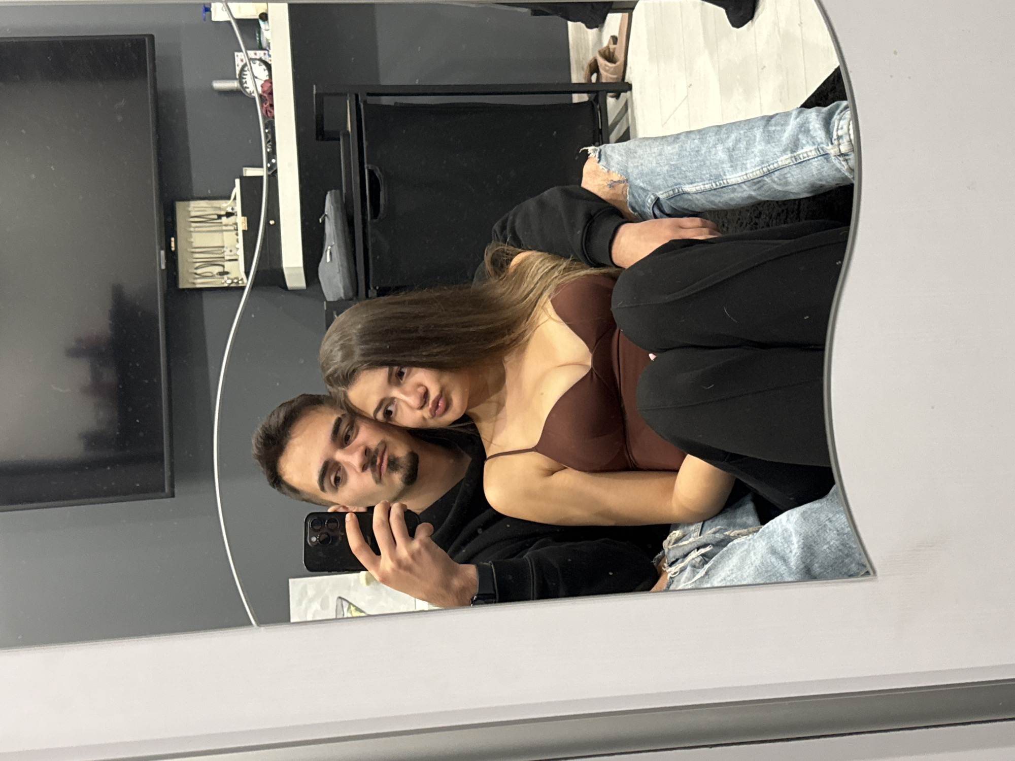 A mirror selfie captures a young couple posing in what appears to be an indoor setting. The image is taken from a reflection in a large, white-framed mirror.
On the left side of the frame, a man with short dark hair and a beard holds a black smartphone, aiming it at the mirror to capture the reflection. He is wearing a black long-sleeved top and a dark watch on his left wrist. He looks directly into the camera with a slight, confident expression. To his right, a woman with long, straight brown hair leans into him. She wears a dark brown or maroon spaghetti-strap top and black trousers or leggings. Her gaze is also directed at the camera, with a subtly pouting expression.
In the background, visible through the mirror's reflection, is a modern room. A large black flat-screen television is mounted on a dark gray wall. Below the TV, a white shelf or storage unit holds various cables and small items, including a small clock with red details. To the right, a black clothing hamper or storage bin and a gray soft case are visible. The floor beneath them appears to be a light-colored wood or laminate. The man's legs, clad in light blue distressed jeans, are visible near the bottom right. The couple appears close and relaxed, capturing a shared moment in what could be a home in Sofia, Bulgaria. No legible text is visible in the image.