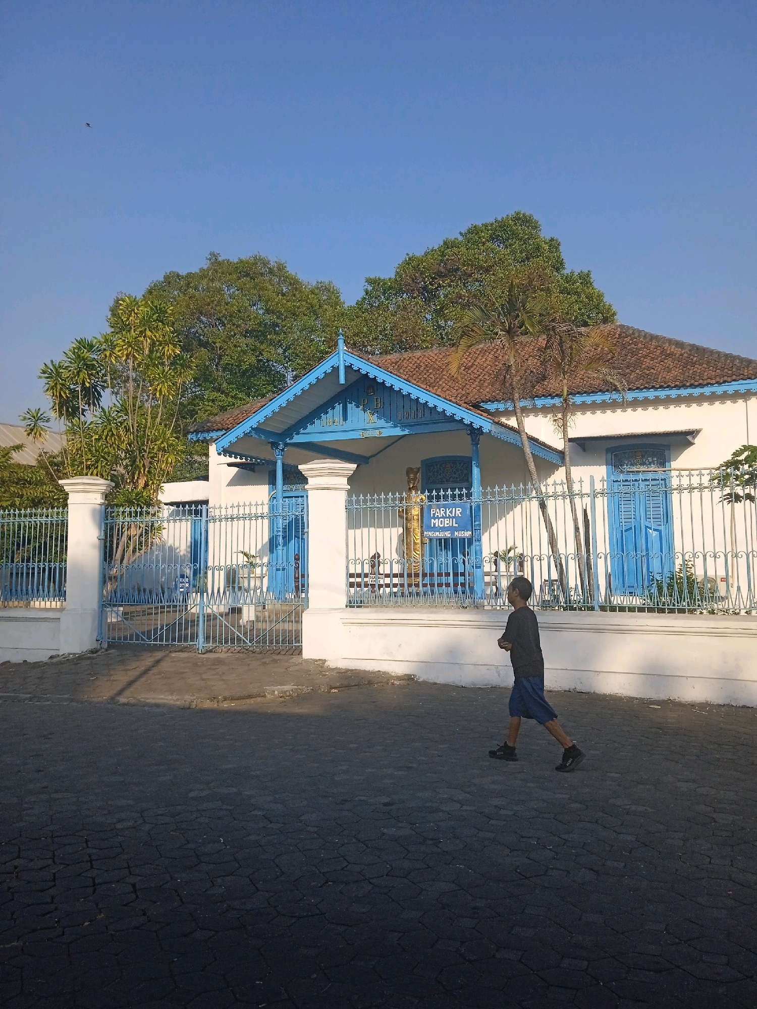 A young boy walks across a paved courtyard in front of a white building with blue accents in Surakarta, Indonesia. The building has a red tile roof and ornate blue trim around the entrance and windows. A blue metal fence encloses the property, and trees surround the building. A sign that reads "PARKIR MOBIL PENGUNJUNG MUSEUM" is visible inside the gate, indicating it is a visitor parking area for a museum. The sky is clear and blue, and the sun casts long shadows, suggesting it is early morning or late afternoon.