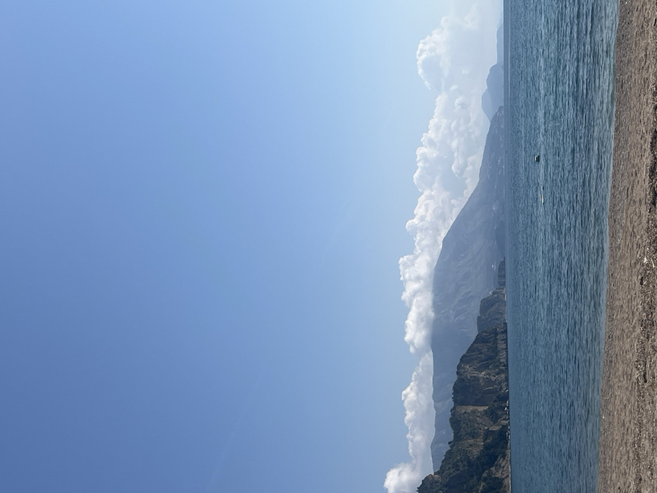 This image captures a panoramic coastal scene from a beach in the city of Unknown, Unknown, under clear daylight conditions.
The foreground consists of a narrow strip of light brown sand, devoid of people or notable objects.
The mid-ground is occupied by a calm body of deep blue-green water, exhibiting subtle surface undulations. A singular dark, cylindrical object, likely a buoy or marker, is visible floating in the water slightly to the right of the frame's vertical center.
The background features a long expanse of rugged coastline defined by a series of imposing, verdant mountains and steep rocky cliffs that extend from the left edge of the frame to the distant right horizon. The upper slopes and peaks of these mountains are partially obscured by a band of dense, white cumulus clouds. Further along the distant horizon to the right, additional smaller landforms or islands are visible, appearing hazy.
The sky, occupying the upper two-thirds of the image, is a consistent, clear light blue with scattered white cumulus clouds concentrated above the mountain range. No people, buildings, or active vessels are present within the entire scene.