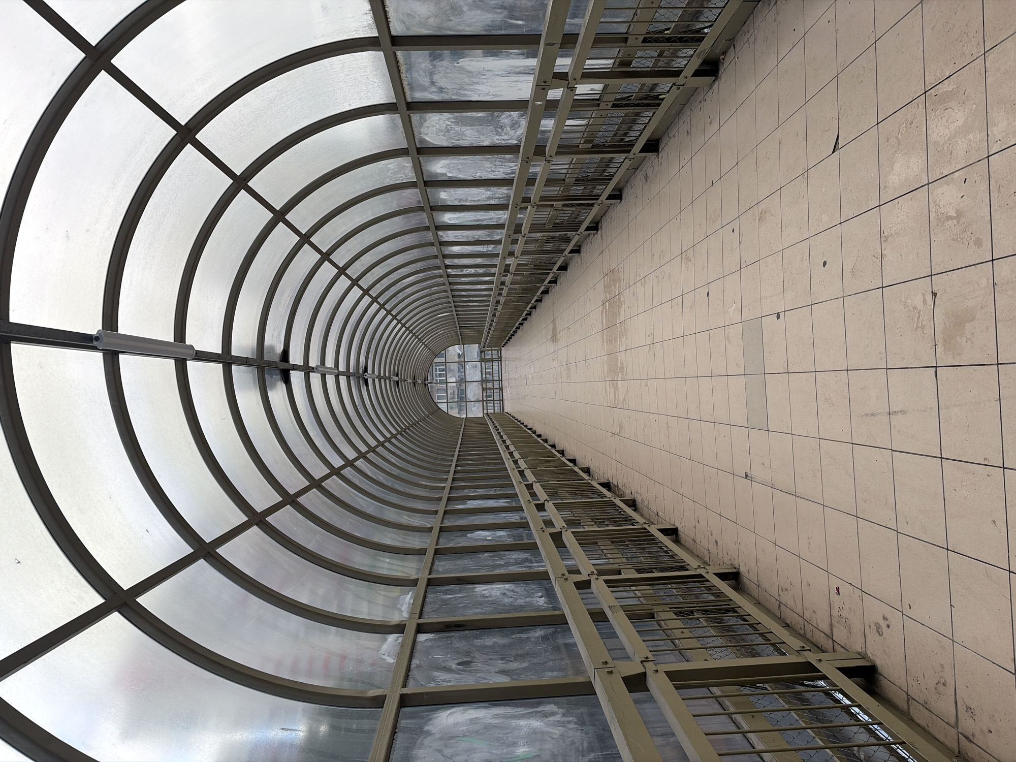 The image shows an enclosed pedestrian walkway in Sochi, Russia. The walkway is constructed with a semi-circular, transparent roof supported by a network of metal frames. Below the roof, a metal structure runs the length of the walkway, parallel to a tiled wall. The tiled wall is made up of beige-colored square tiles. The perspective suggests the photo was taken at a low angle, looking up the length of the walkway. The end of the walkway is visible in the distance.