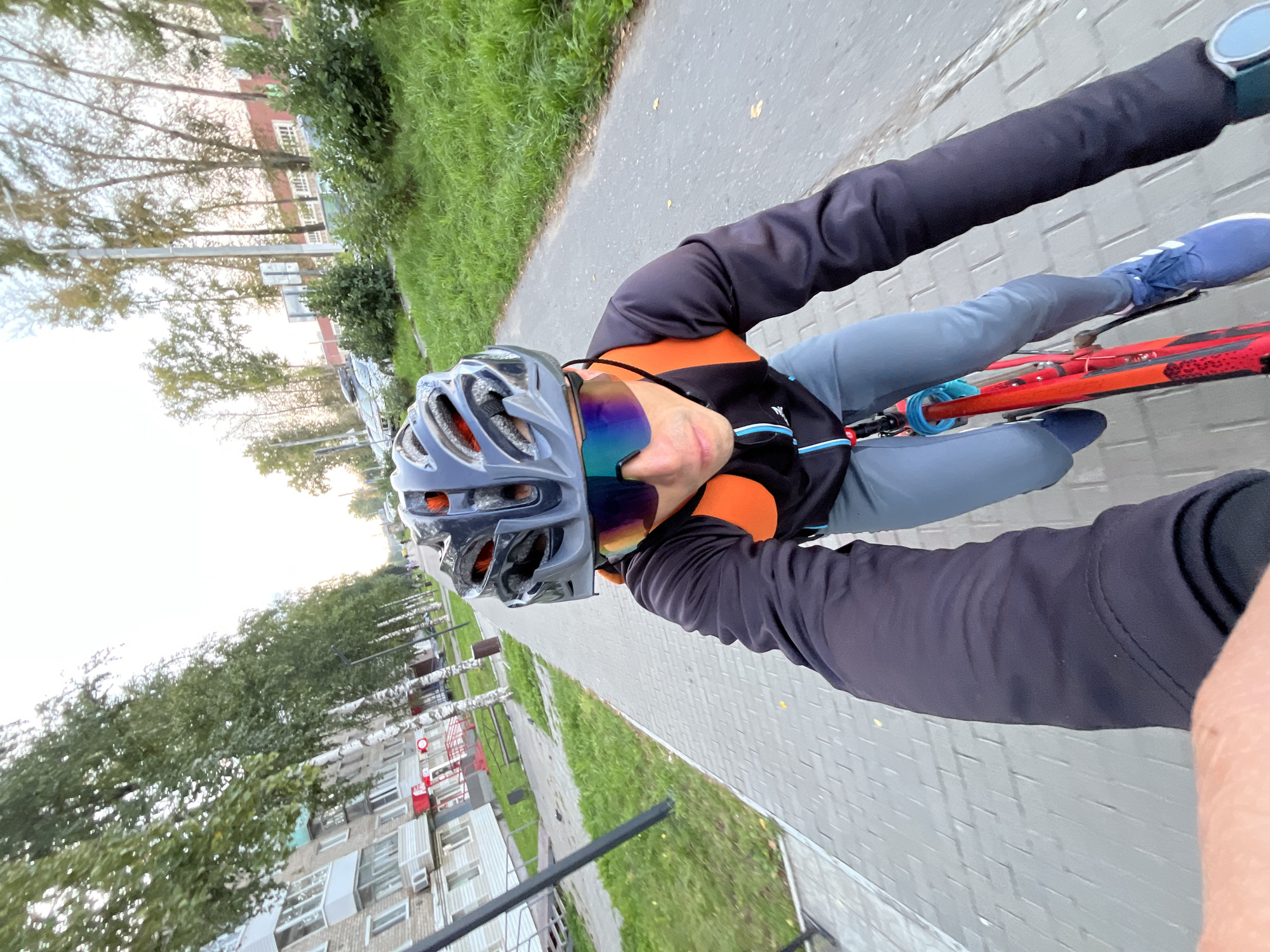 A first-person perspective shot shows a person riding a bicycle on a paved path in Chaikovsky, Russia. The rider is wearing a grey helmet and reflective sunglasses. They are dressed in a black cycling jacket with orange accents, light grey cycling pants, and blue athletic shoes. The bicycle is red. The path is lined with green grass and trees, with apartment buildings visible on the left side of the frame and more trees and buildings on the right. The sky is overcast.