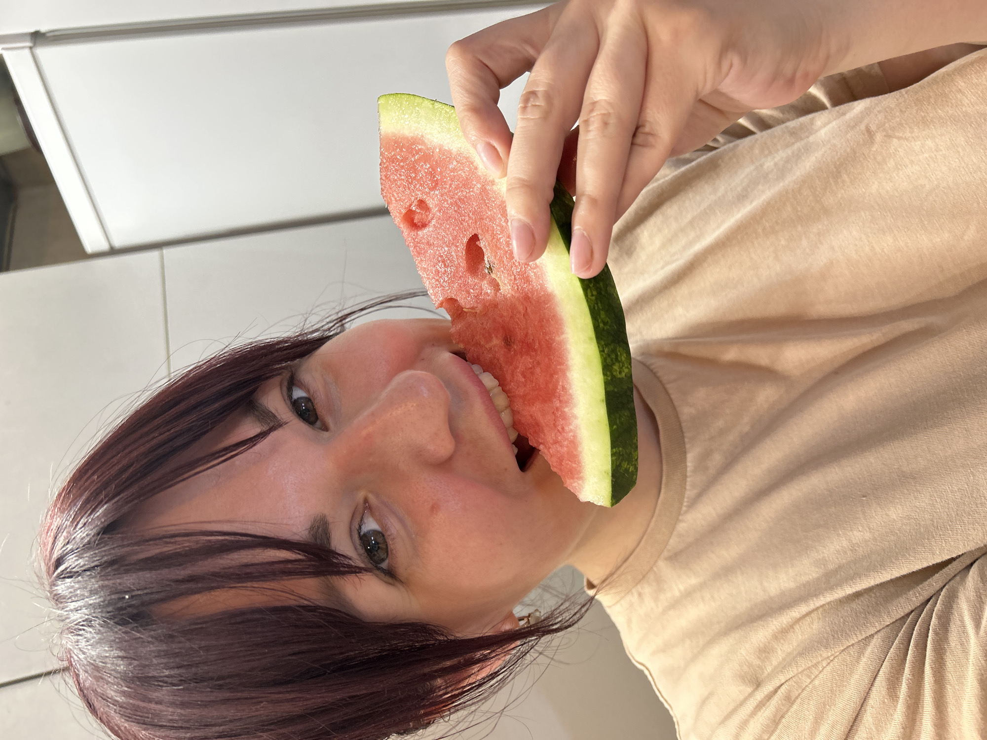A young woman with dark reddish-brown hair and bangs is taking a bite out of a slice of watermelon. She is holding the watermelon in her right hand, and a small piece is already bitten off. Her left hand is not visible. She is wearing a light beige t-shirt. The background appears to be an indoor setting, possibly a kitchen, with white cabinets visible on the right. The lighting suggests an artificial light source, likely from above. The image is a close-up shot, focusing on the woman's face and the watermelon. There is no indication of the location in the city of Фоки, Russia.