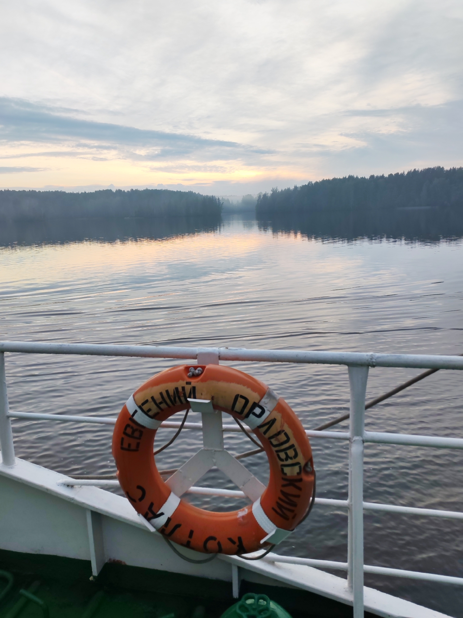 The image shows a life preserver attached to a railing on the deck of a boat. The life preserver is orange with white stripes and has Cyrillic text on it. The boat is on a body of water, likely a river or lake, with a tree-lined shore in the distance. The sky is cloudy and overcast, with the sun breaking through in the distance. The scene appears to be set in the early morning or late evening. The location is identified as Kurikaevo, Russia.