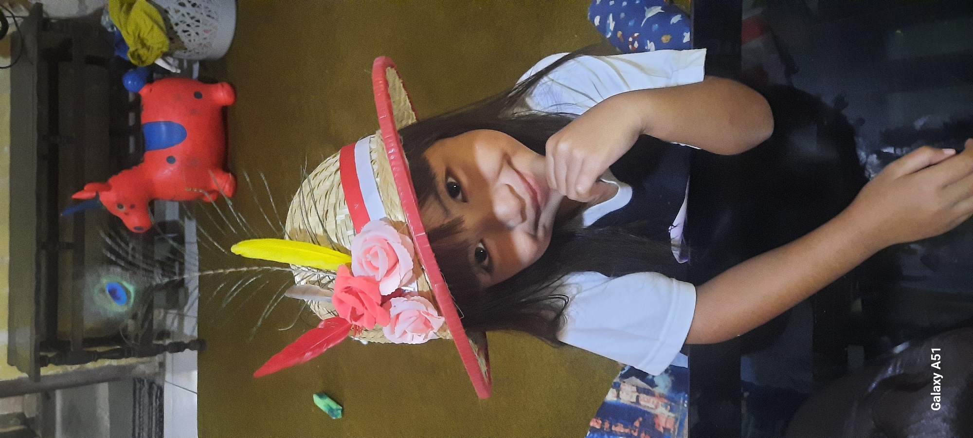 In Jatitengah, Indonesia, a young girl with long, dark hair is seated, wearing a straw hat adorned with pink roses and red and yellow feathers. She is dressed in a white t-shirt over a dark garment. Her right hand is raised to her chin, and she is smiling at the camera. In the background, a red inflatable toy horse with blue accents is visible, along with a peacock feather. The setting appears to be indoors, with a wooden furniture piece and a white laundry basket in the background. The overall lighting is dim.