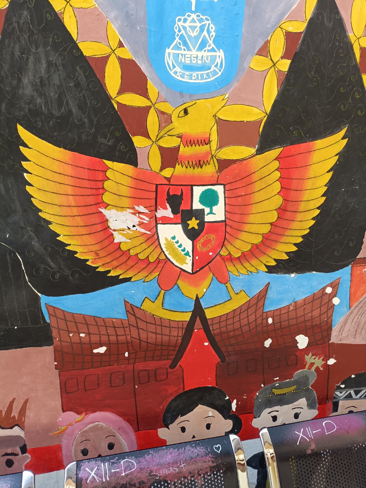 The image displays a colorful mural, likely painted on a wall, featuring symbolic elements and stylized figures. Prominently featured is the Garuda Pancasila, Indonesia's national emblem, depicted with outstretched wings in shades of yellow and red, holding a shield with various symbols representing the Pancasila principles. Above the Garuda, a blue crest with white lettering reads "NEGERI KEDIRI," indicating its connection to Kediri City.
Beneath the Garuda is a depiction of traditional Indonesian architecture, possibly a rumah adat or a stylized representation of buildings. The lower portion of the mural shows stylized heads of several children, each with distinct features and hairstyles. One child wears a pink headscarf, another has a simple black haircut, and a third has dark hair adorned with a golden band and a sprig of greenery. The children are positioned as if peeking over a surface that has "XII-D" and some handwritten text on it. The overall artwork is vibrant and appears to be a community or school project. The mural's style is naive and decorative, with bold outlines and flat colors. The background features abstract patterns in black, brown, and yellow.
Given the presence of "NEGERI KEDIRI" and the nature of the artwork, it is highly probable that this mural is located in a public space within Kediri City, Indonesia, such as a school, community center, or a government building.