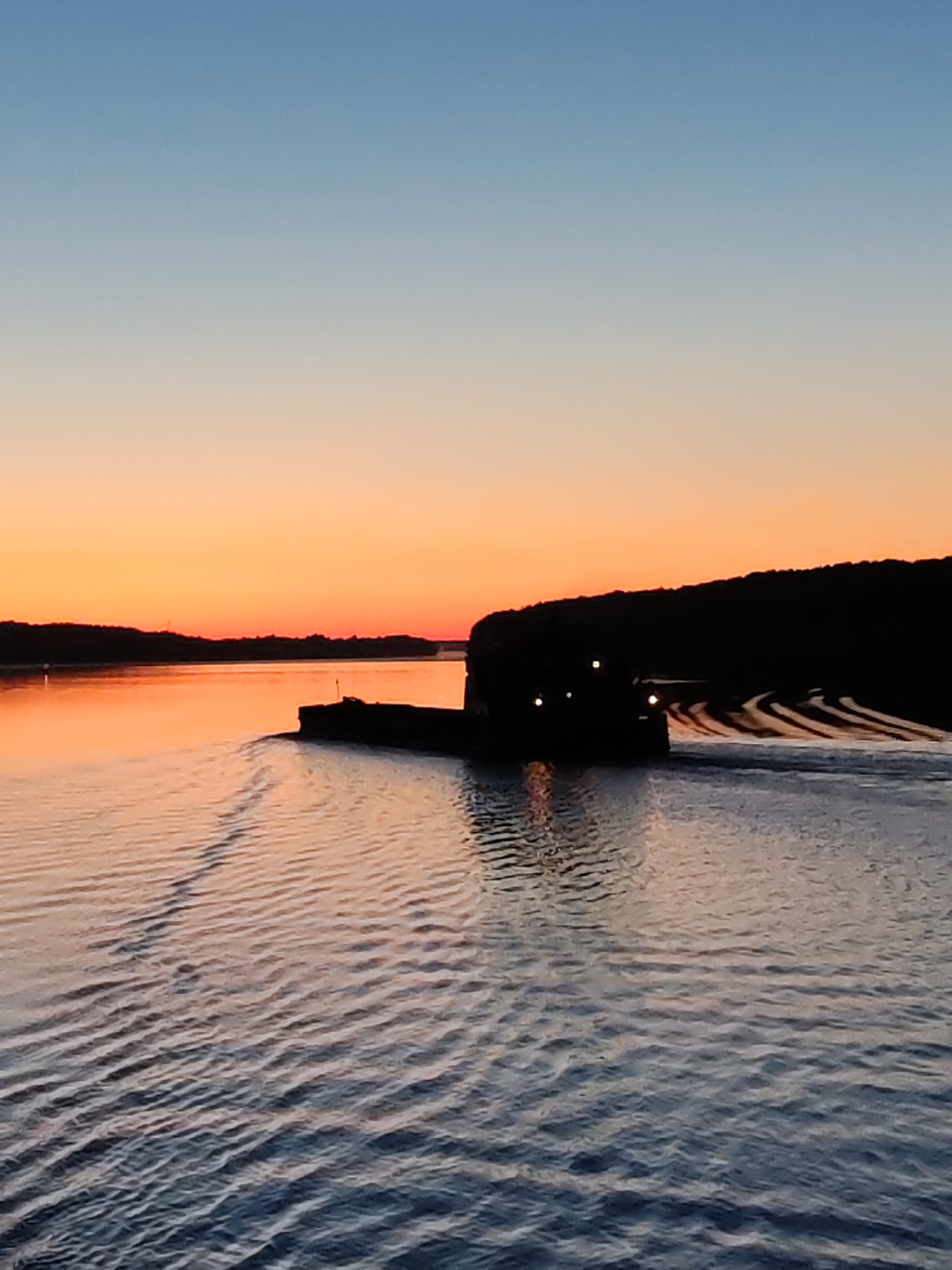 The image, set in the context of Раменье, Russia, depicts a serene twilight scene over a wide body of water, likely a river or large lake. The sky presents a distinct gradient, transitioning from a light, clear blue at the top to a vibrant orange and deep red at the horizon, indicative of either sunrise or sunset, with no visible clouds. This warm light is reflected on the water's surface, creating a luminous orange glow closer to the distant shorelines, which then fades into cooler shades of blue and grey towards the foreground.
A prominent, dark-silhouetted vessel, appearing to be a barge or a tugboat pushing a barge, occupies the mid-ground, positioned slightly to the right of the frame. Several small, bright yellow-white lights are visible on the vessel's structure, contrasting sharply with its overall dark form against the colorful sky. The vessel is actively moving from left to right, creating a distinct, V-shaped wake that trails behind it, forming parallel ripple patterns on the water's surface.
On the left side of the horizon, a faint, dark, indistinct shoreline is visible against the glowing sky. To the right, a more substantial landmass, heavily silhouetted and appearing as a dark, irregular line, likely a forested bank or hillside, extends along the horizon. The foreground water is characterized by numerous small, undulating ripples, which reflect the ambient light and provide texture to the scene. No individuals are discernible within the frame.