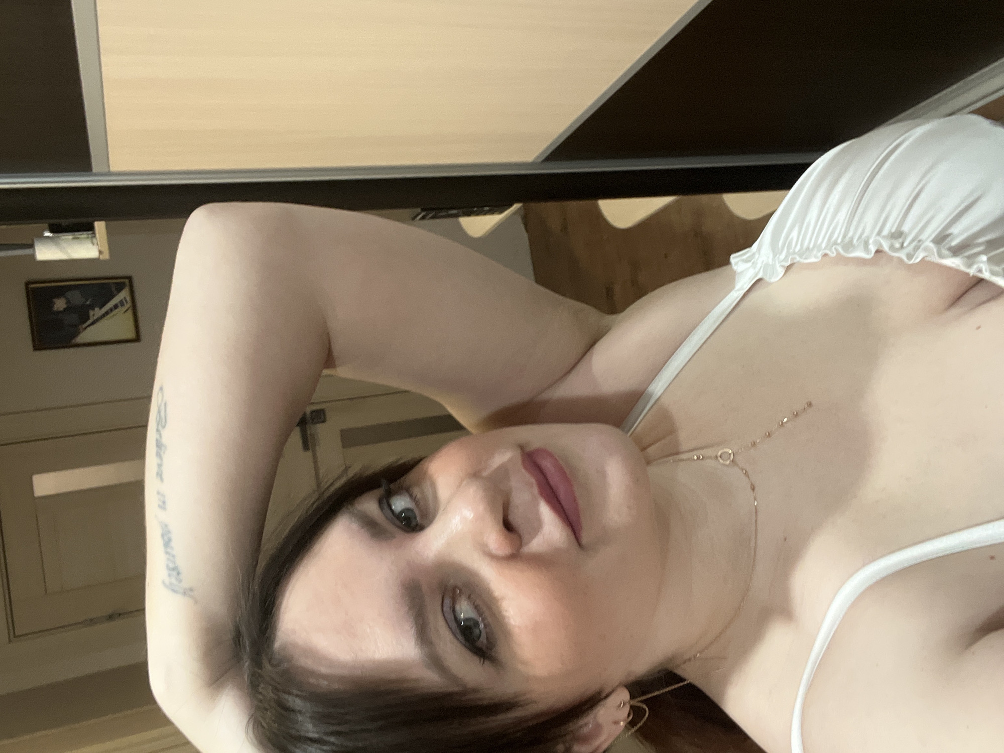 A fair-skinned woman with dark hair, wearing a white top with thin straps, lies on her back. Her left arm is raised above her head, revealing a tattoo on her inner forearm. She is looking directly at the camera with a neutral expression. A gold necklace is visible around her neck. The background includes a wall with a framed picture and part of a closet. The location in the city of Noviy, Russia, is not discernible from the image.