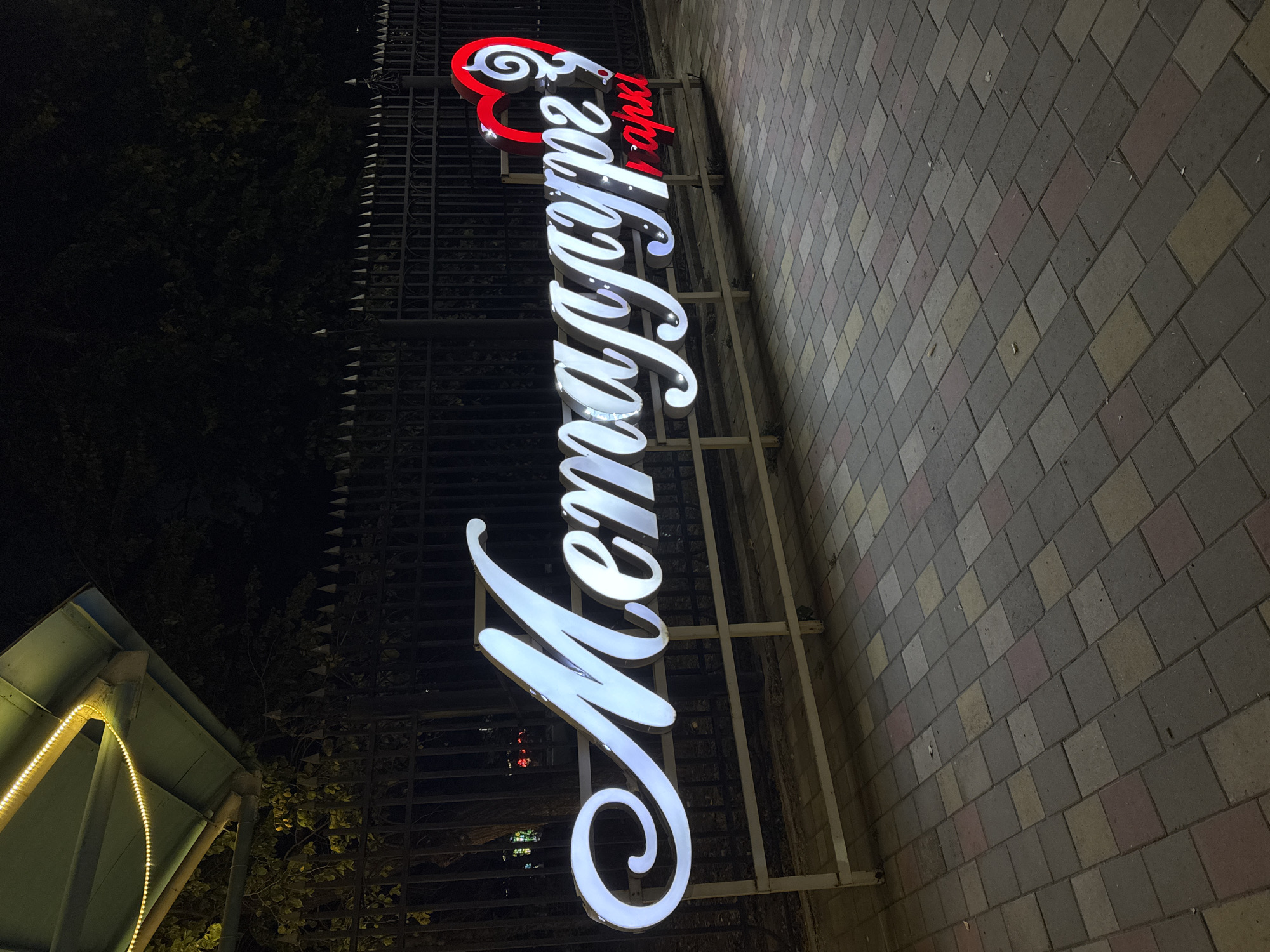 An illuminated sign for "Metallurg Park" is displayed at night in Oskemen, Kazakhstan. The sign, in Cyrillic script, features white, cursive lettering with internal lights, and a red heart symbol on the right side. The sign is mounted on a white metal frame and stands in front of a black metal fence. The ground is paved with interlocking bricks in various shades of grey, pink, and yellow. Trees and darkness are visible behind the fence, and a section of an illuminated canopy with yellow lights is visible in the upper left corner of the image.