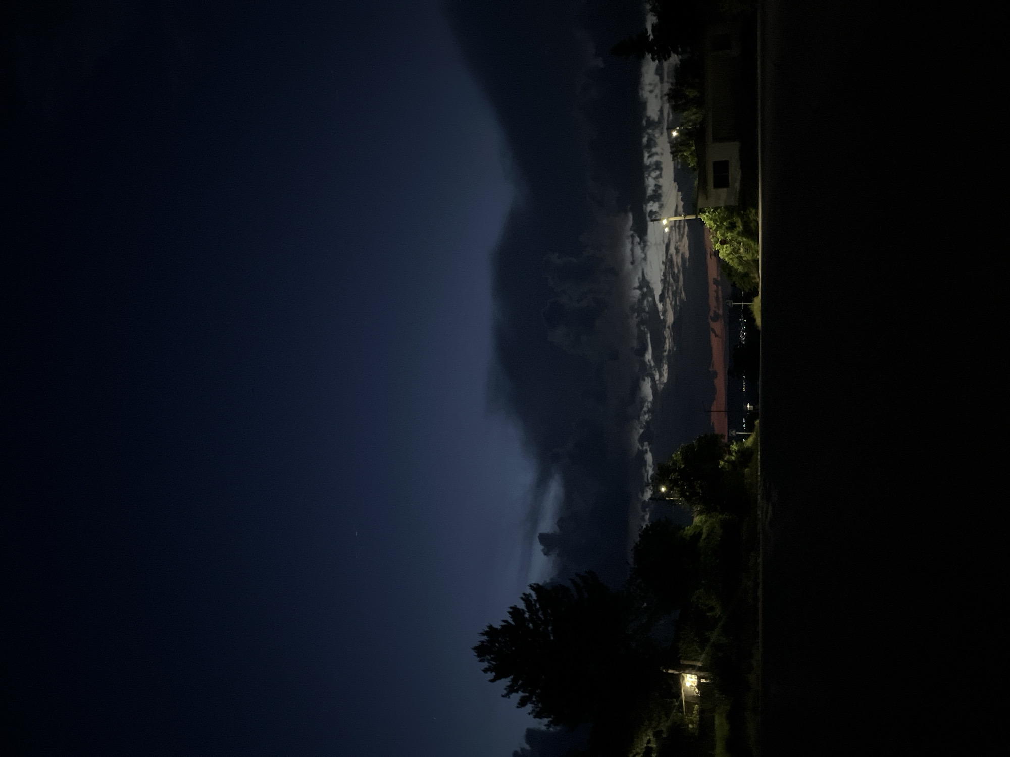 The image depicts a nighttime scene in Ivanski, Bulgaria, characterized by deep twilight and artificial illumination. The upper two-thirds of the frame are dominated by a dark indigo to navy blue sky, transitioning to lighter blues at the horizon. Prominent, dense, dark gray to black cumulus clouds traverse the mid-sky horizontally, with more fragmented, lighter gray clouds visible beneath them. A faint reddish-orange glow is present along the distant horizon. Scattered, minute white points, consistent with stars, are visible in the clearer sky sections.
The foreground, comprising the bottom third of the image, is a uniformly dark, unlit surface, likely an asphalt road or ground.
In the midground, on the left, a large, dark silhouette of a coniferous tree, likely a pine, stands prominently. Below this tree, a single-story building is partially visible, with a distinct warm yellow light source emanating from an interior opening or window. Further right, two street lights with bright, warm yellow illumination are visible; one illuminates a small, partially obscured building and adjacent foliage on the right side, while another is positioned further back on the left side, its light partially hidden.
The distant horizon features a subtle outline of undulating terrain or structures. On the far left of the horizon, a cluster of faint, twinkling yellow-orange lights indicates a distant settlement or city. A light-colored rectangular sign on a pole is barely discernible on the left side of the road, adjacent to the distant lights.
No human figures or explicit actions are discernible within the frame. The scene is illuminated by natural twilight and localized artificial street and interior lighting, resulting in deep shadows.
