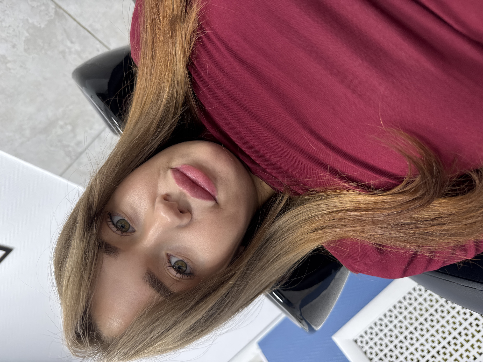 A young woman with shoulder-length, light brown hair and green eyes is captured in a close-up, head-and-shoulders selfie. She is wearing a dark red, V-neck top, and her lips are painted a light pink. Her gaze is directed towards the camera. The background shows a portion of a black chair, a blue wall, and a white decorative grate. The overall lighting is bright and even. This photograph appears to have been taken indoors, likely in a salon or personal care establishment. The specific location in Chaikovsky, Russia is not discernible from the visual elements of the image.