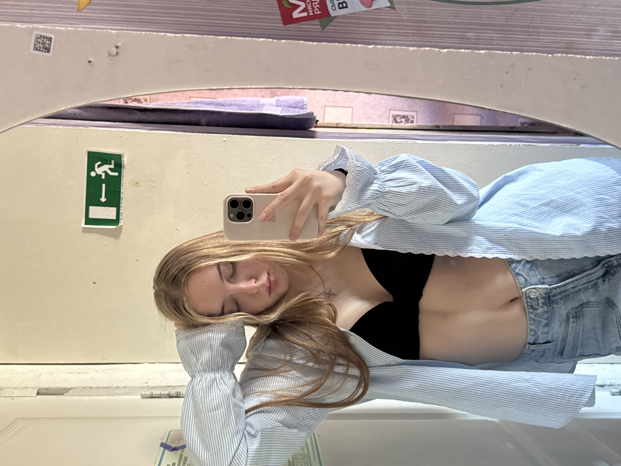 A young woman is taking a selfie in what appears to be a public restroom. She is holding a smartphone in her right hand, capturing her reflection in the mirror. She is wearing a black bra, a blue and white striped blouse, and light blue jeans. Her blond hair is loose around her face and shoulders. The background shows a typical public restroom interior with white tiles. A green exit sign is visible on the wall. A red snack package is visible on the shelf above. The city mentioned in the image request, Perm, Russia, cannot be verified from the image.