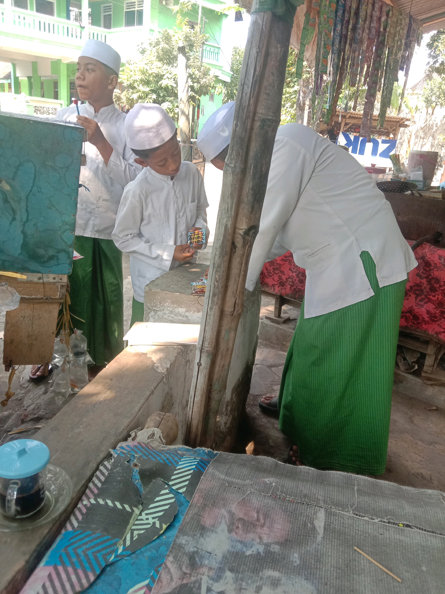In the city of Ngempit, Indonesia, three young males, dressed in traditional white Islamic attire and white caps, are gathered around a makeshift stall. One individual, wearing a green sarong, is leaning over, his back to the camera, holding what appears to be a small object. Another boy in a white shirt and green sarong is focused on a small, colorful item he holds in his hand. The third boy, also in white, stands slightly behind them, looking towards the left.
The stall itself is constructed from rough wood and stone. Bags of what appear to be fish or shrimp, in plastic packaging, are suspended from the side of the stall. In the foreground, a glass of dark liquid, possibly coffee, sits on a saucer next to a worn, patterned mat. The background reveals a green building with multiple windows and trees, suggesting an outdoor setting, possibly near a school or religious institution. The overall scene depicts a casual interaction at a small roadside stall.