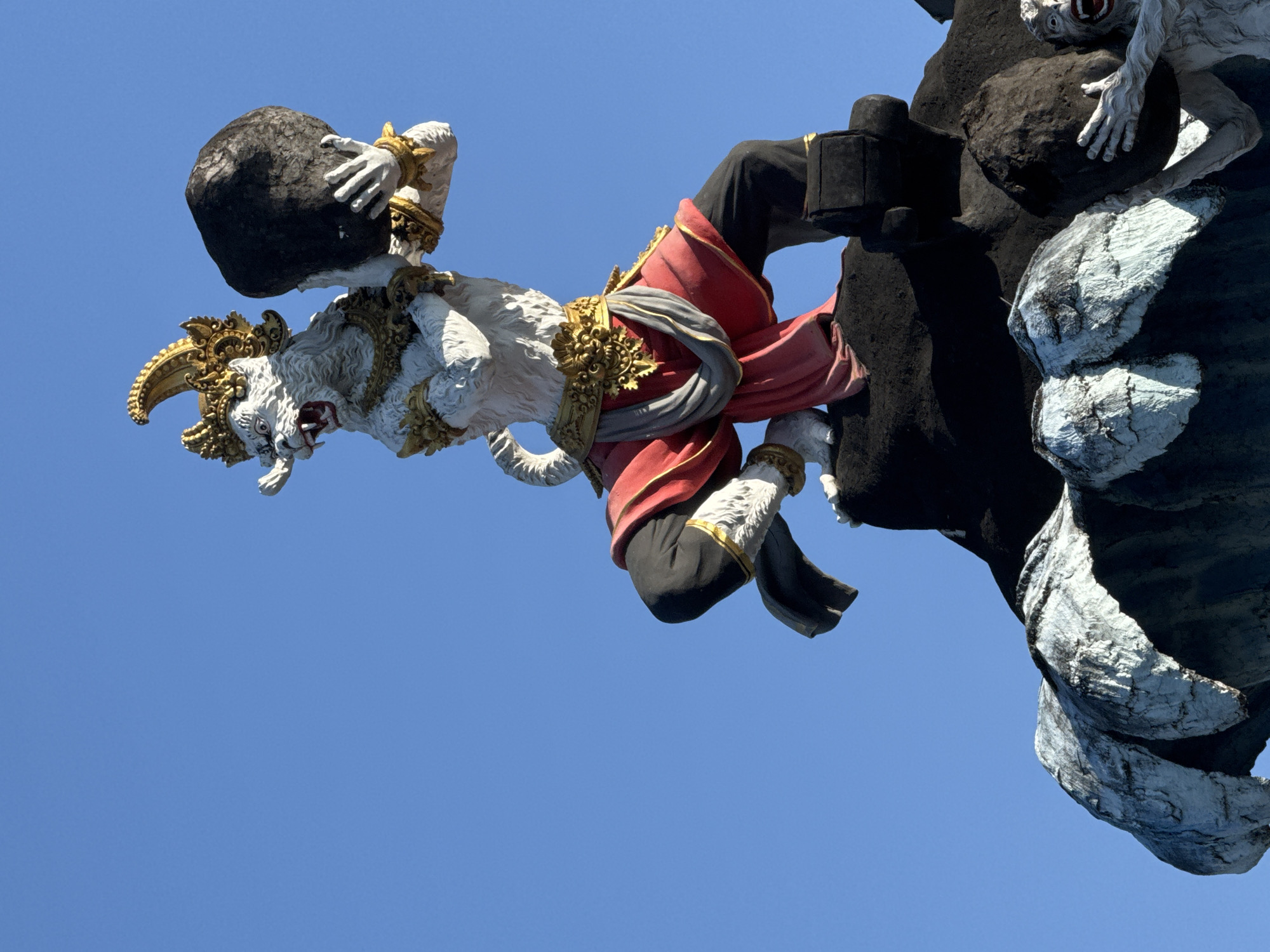 A low-angle shot of a Balinese statue in Denpasar, Indonesia, shows a white mythical creature, possibly a Rakshasa demon, sculpted with golden headdress and ornate golden shoulder and waist decorations. It's carrying a large black boulder with both hands. The demon is part of a larger, dark gray, boulder-like structure from which another lighter gray, human-like figure emerges at the top right. The demon is wearing a red garment with a gray sash. The background is a clear, bright blue sky. The statue appears to be part of a larger construction, like a temple or a gate, given the placement of the statue.