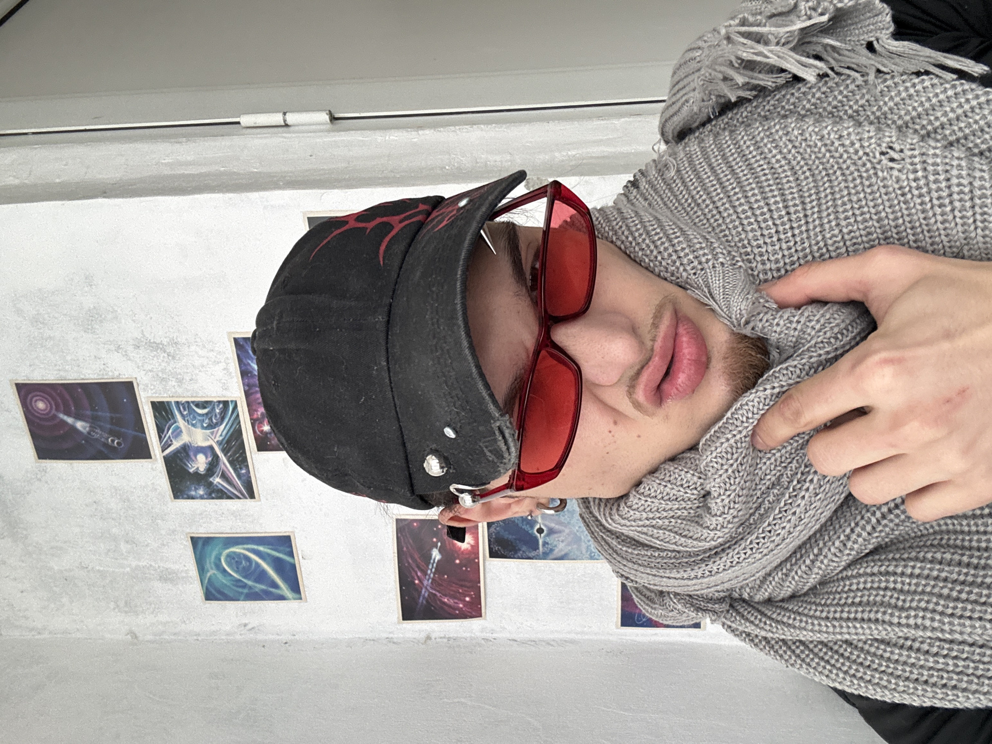 A person is wearing a black baseball cap with red designs, red-tinted sunglasses, and a grey scarf. The person is holding the scarf around their neck with their right hand. The background features a white wall adorned with small, framed images of celestial or cosmic designs. The exact location in Sofia, Bulgaria, cannot be determined from this image.