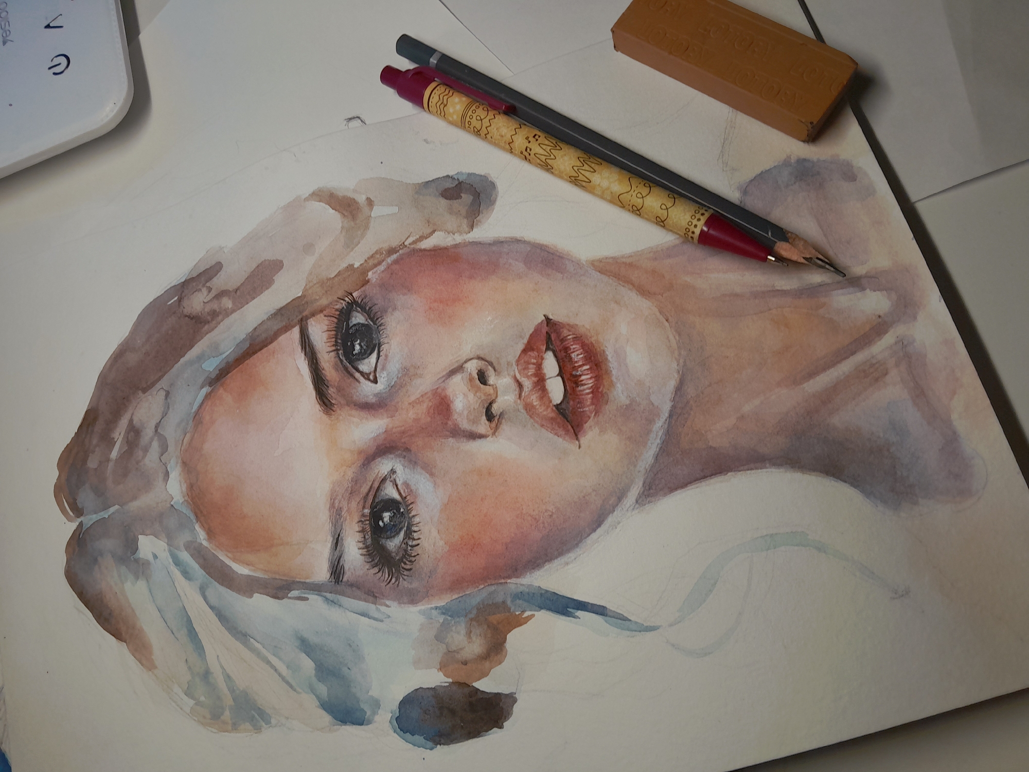 The image shows a close-up watercolor painting of a woman's face on white paper. The painting is oriented at an angle, with the top of the head to the left. The woman has fair skin with rosy cheeks, detailed eyes with long lashes, and full red lips. Her hair appears to be light brown or blonde, with hints of blue in the painted strands. The background of the painting is unfinished.
In the upper right of the image, there is a rectangular brown eraser with the word "VICTORY" embossed on it. Two pencils are positioned over the painting: one is a dark grey pencil, and the other is a lighter colored pencil with a decorative pattern and a red tip. In the upper left corner, a portion of a white object, possibly a digital drawing tablet, is visible. The scene is illuminated by overhead lighting, casting soft shadows.
There is no visible connection to Tashkent, Uzbekistan, in this image.