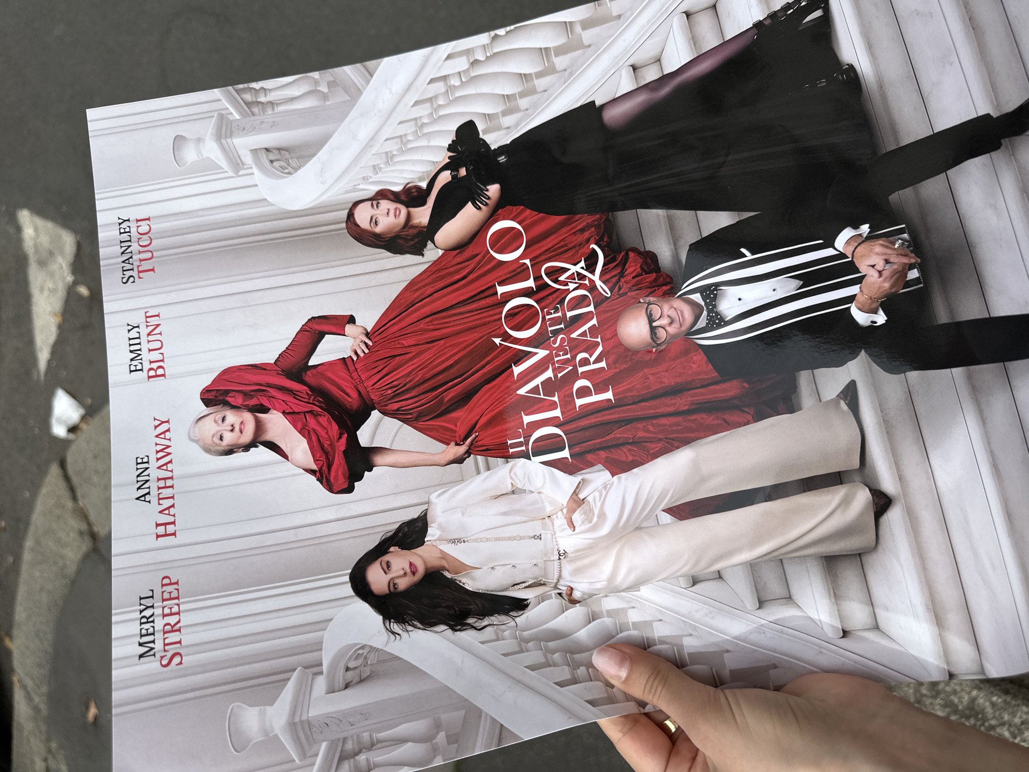 A person is holding a glossy poster outside, which displays the cast of "The Devil Wears Prada 2" on a white balustrade staircase. From the top to bottom, the cast members are Stanley Tucci in a black and white striped tuxedo, Emily Blunt in a red gown and black gloves, Anne Hathaway in a white pantsuit, and Meryl Streep in a red dress. The title, "Diavolo Veste Prada 2," is superimposed over the image. The location is Milan, Italy.