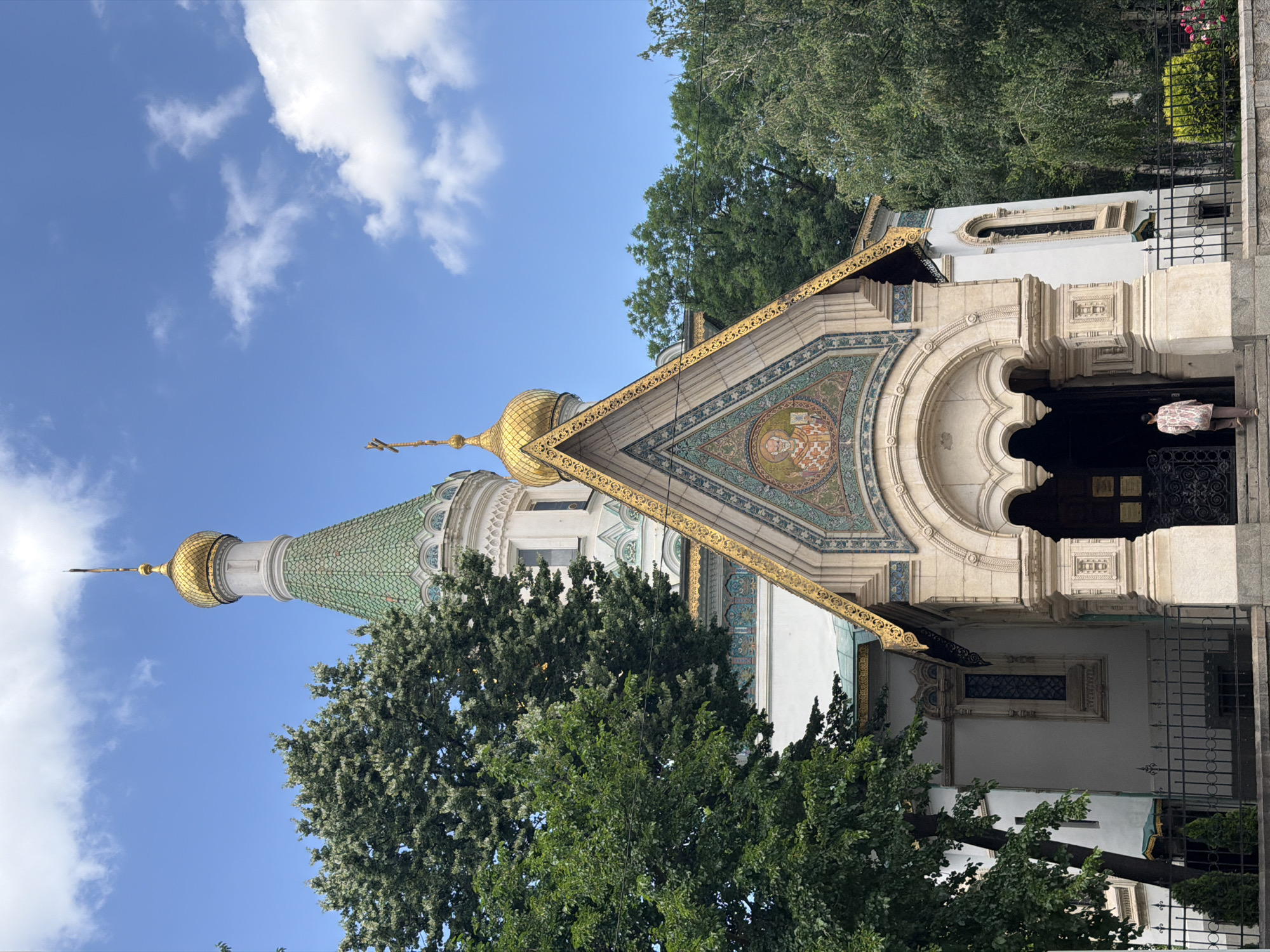 This vertical image depicts the ornate facade and towers of the St. Nicholas the Wonderworker Russian Church in Sofia, Bulgaria, under a bright blue sky dotted with scattered white clouds. The church exhibits a distinct Russian Revival architectural style, characterized by its vibrant colors and intricate details.
The central part of the image focuses on the main entrance, which is crowned by a steeply pitched roof with golden trim. Below this golden roof is an elaborate mosaic panel, featuring religious figures and detailed patterns. The main entrance itself is an arched dark doorway. To the left, a tall tower rises, topped with an emerald green tiled conical roof, which culminates in a smaller golden onion dome and a spire. Another golden onion dome is visible on a shorter tower to the right, also topped with a spire and an Orthodox cross.
Lush green trees with dense foliage partially obscure the church's lower sections and frame it on either side, suggesting a park-like or garden setting. Bright sunlight illuminates the scene, making the golden domes and decorative elements gleam. Two individuals are visible near the main entrance; one is largely obscured by the shadow of the doorway, while the other, wearing a patterned shirt, appears to be approaching or leaving the entrance. The bright lighting and clear sky suggest it is daytime, likely morning or afternoon, conveying a peaceful atmosphere. No clearly legible text is discernible on the church facade.