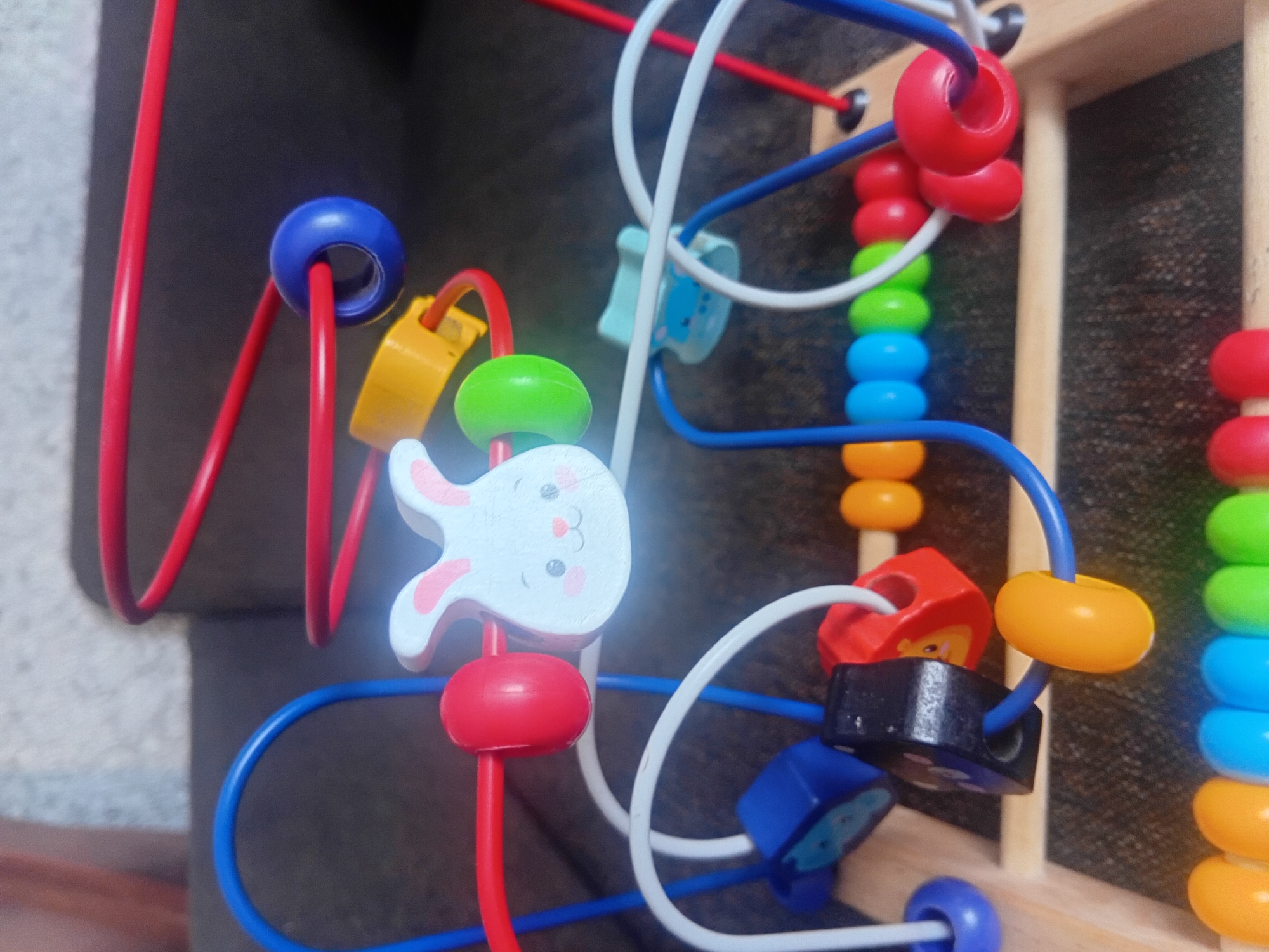 This is a close-up, eye-level, indoor photograph of a colorful wooden bead maze toy. The toy is primarily made of natural wood with various colored beads and plastic components. The beads are made of wood and plastic in bright colors, including red, blue, yellow, green, and orange. Some of the beads are round, while others are shaped like animals or blocks with graphics.
The main subjects are the components of the bead maze. A prominent feature is a white wooden rabbit with pink accents on its ears and cheeks. This rabbit is positioned on a red plastic bead. Other notable objects include red, blue, and yellow plastic wire loops that guide the beads, and a stack of colorful wooden beads on the right side of the frame, arranged vertically in a rainbow sequence. There are also several wooden blocks with simple graphics, including one that appears to be a black cube with white circles and a blue block with an image of a bear.
The background is dark and out of focus, suggesting an indoor setting, possibly a room with a sofa or upholstered furniture. The lighting appears to be artificial, casting some highlights on the toy's surfaces. There are no visible people, and the activity taking place is implied by the nature of the toy, which is designed for manipulation and play, likely by a child. No text is visible in the image. The overall impression is one of a playful, engaging children's toy.
