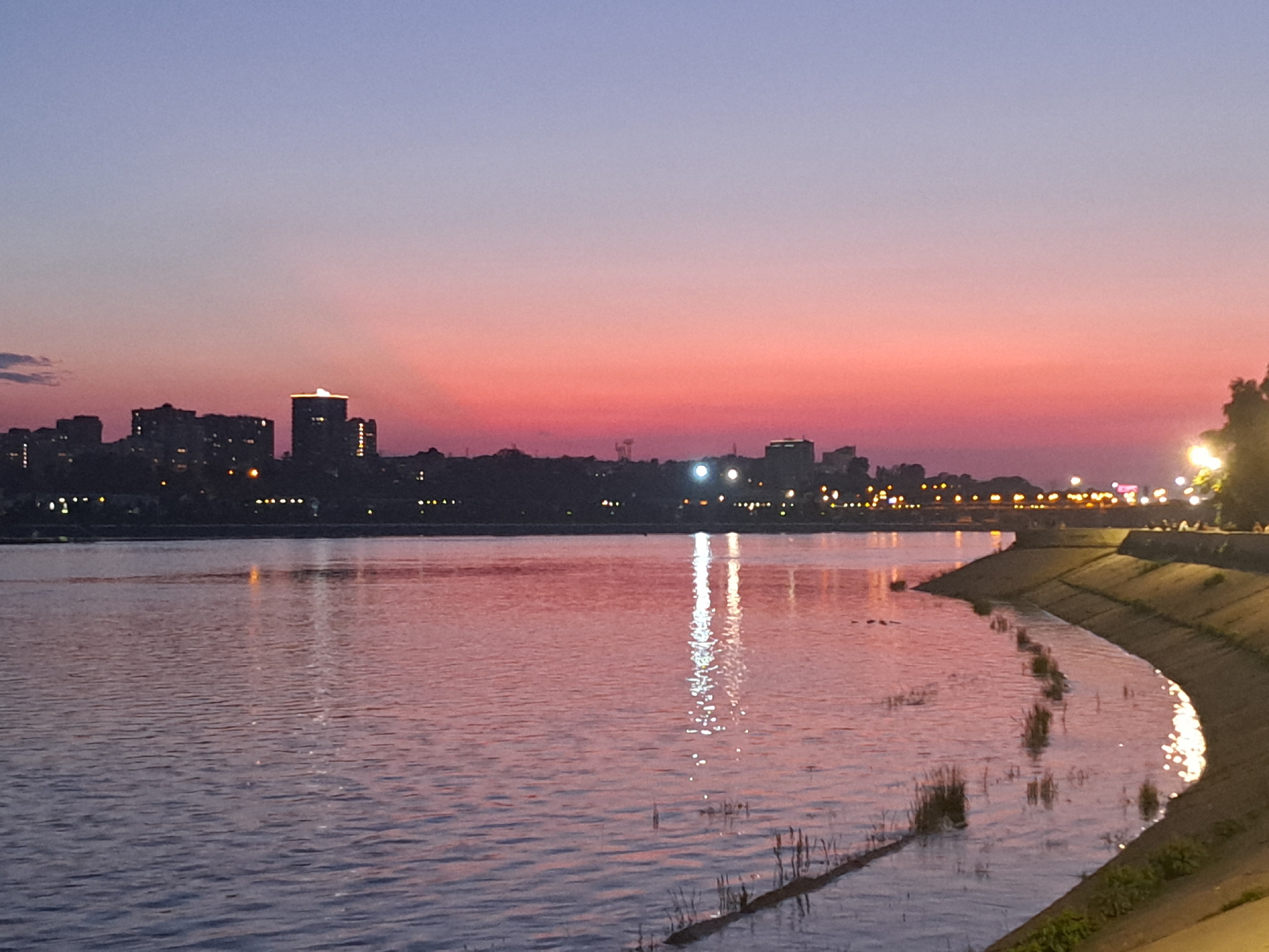 This is a wide-angle, eye-level shot of the Angara River at dusk in Irkutsk, Russia. The sky is a gradient of soft purple at the top fading into a vibrant pink and orange sunset closer to the horizon. A silhouetted cityscape stretches across the background, with a cluster of buildings and a few taller structures illuminated by artificial lights. The water in the foreground is calm, reflecting the colors of the sky and the distant city lights, creating a shimmering effect. A concrete embankment lines the right side of the river, with some sparse vegetation growing along its edge. The overall atmosphere is serene and peaceful.