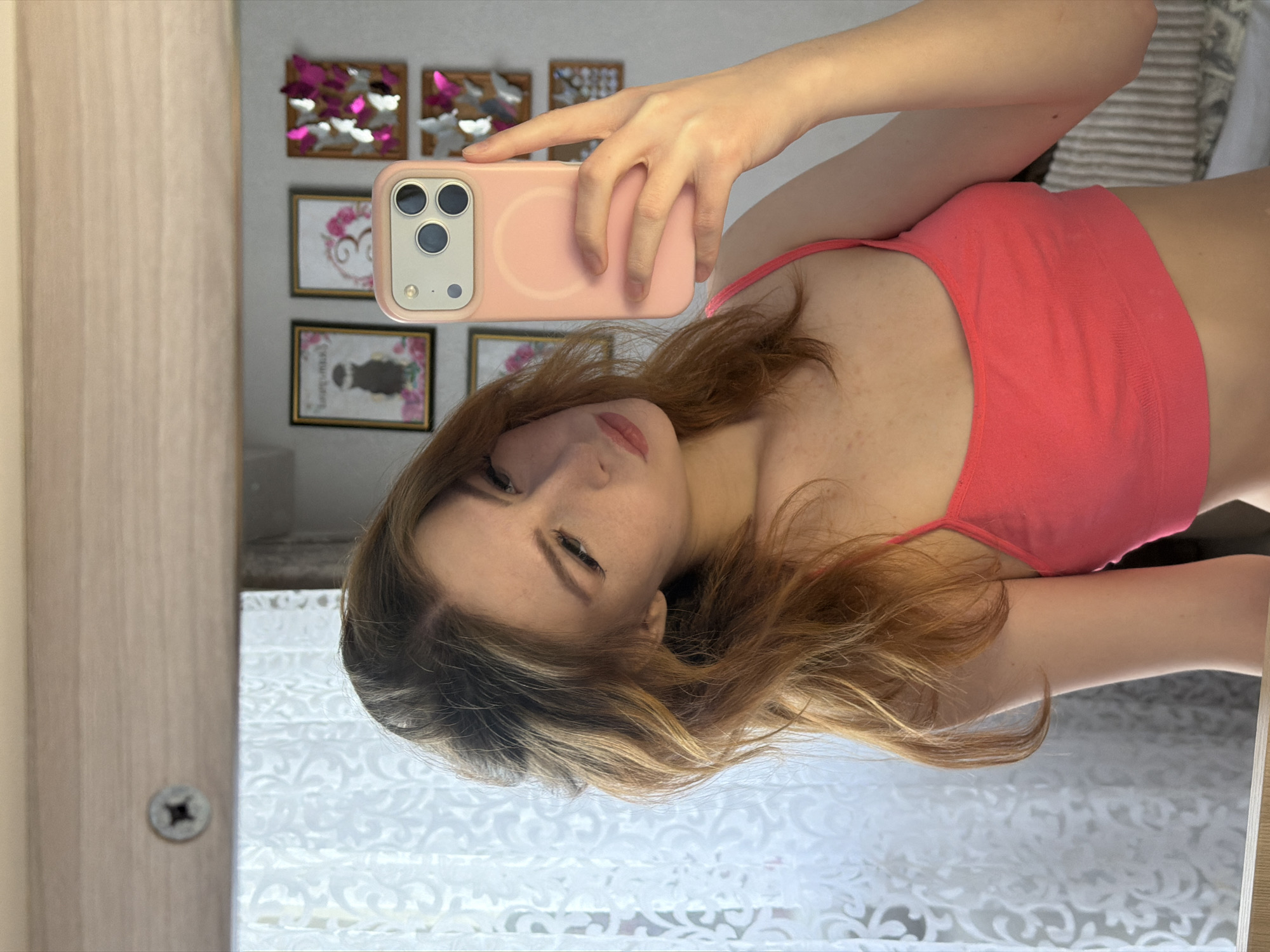 A fair-skinned young woman is taking a selfie in a mirror. She's wearing a pink spaghetti-strap tank top. Her hair is light brown with blonde highlights. She is holding a pink iPhone, partially obscuring her face. In the background, there are framed pictures on the wall with floral and butterfly motifs. There is a white patterned curtain, a light-colored wooden door frame, and a portion of a striped cushion or pillow visible. The image does not provide any information about the location being in the city of Noviy, Russia.