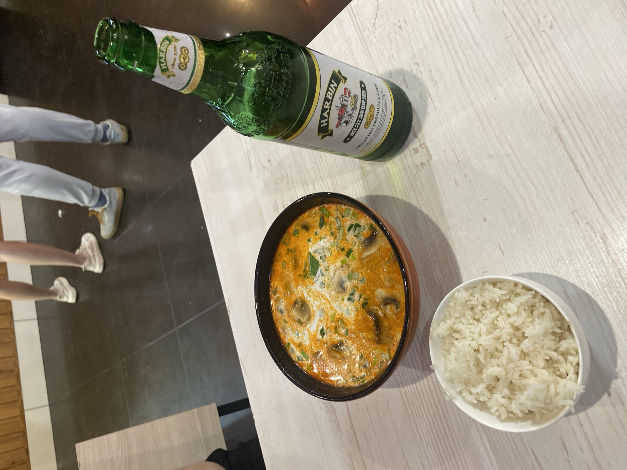 A top-down, slightly angled shot shows a table setting with a bottle of Harbin beer, a bowl of soup, and a bowl of rice. The soup appears to be a creamy, orange-colored broth with mushrooms and green onions. The rice is white and fluffy in a small white bowl. The beer bottle is green with a white label. The table has a light-colored, wood-grain surface. In the periphery, sections of flooring, light blue jeans, and shoes are visible. The location is likely in a restaurant or dining establishment in Krasnoyarsk, Russia, as Harbin beer is imported from China and widely available in the city.