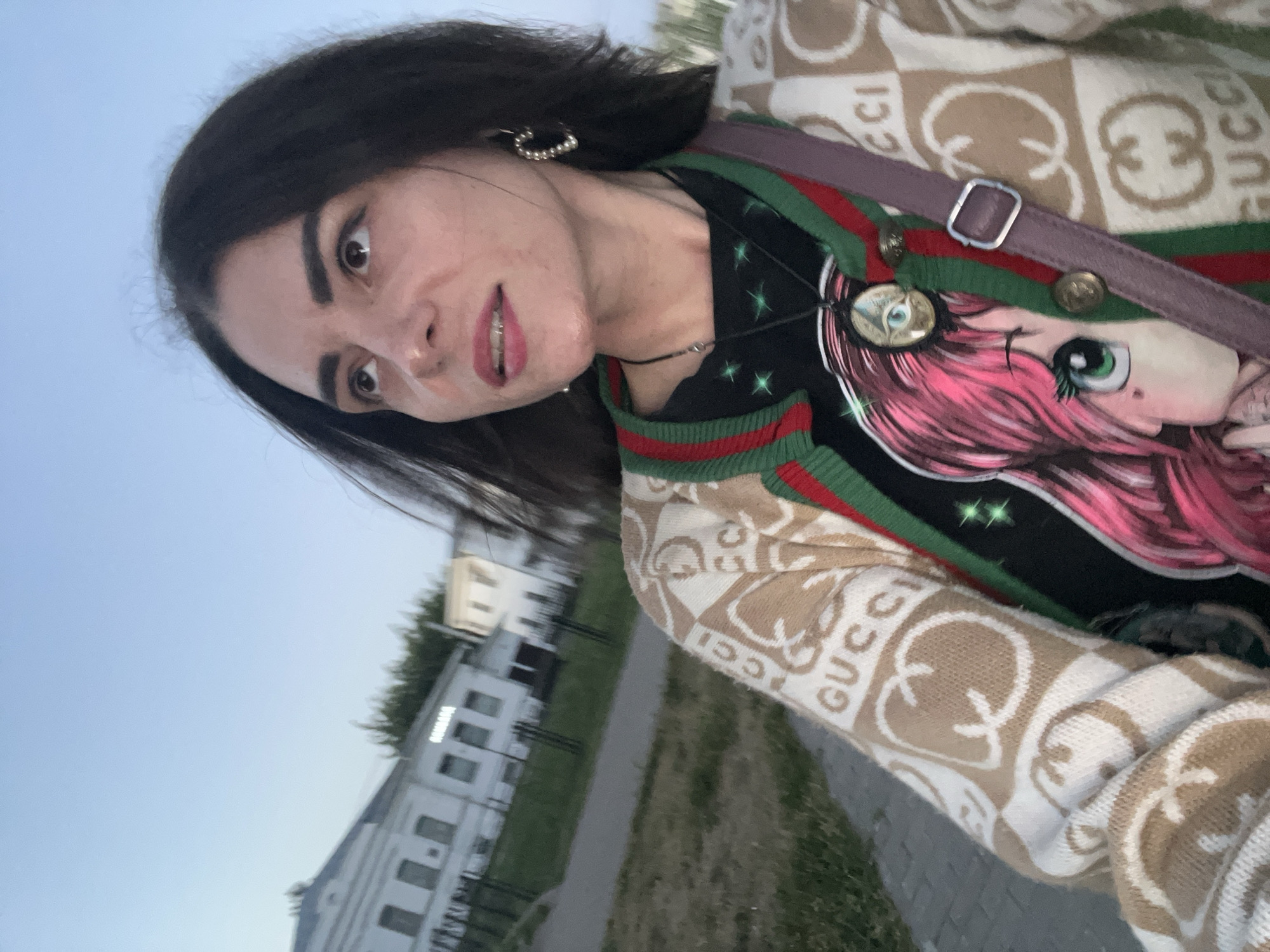 This is a tilted, candid, medium shot of a young woman outdoors. She has dark hair, dark eyebrows, and is wearing pink lipstick and pearl earrings. She is looking directly at the camera with a neutral expression.
She is wearing a black t-shirt with a cartoon character design and a beige cardigan with a repeating "GUCCI" logo pattern. A purple strap from a bag is visible across her chest.
The background shows a building with windows and trees, suggesting an urban or suburban setting. The sky is a pale blue, indicating it's likely daytime, possibly late afternoon or early evening. The ground appears to be a sidewalk next to a grassy area.
There is no specific activity or event taking place; it appears to be a casual selfie.
Visible text: "GUCCI" is prominently visible on the woman's cardigan. A word is also visible on the building in the background, which reads "КОНТАКТ".