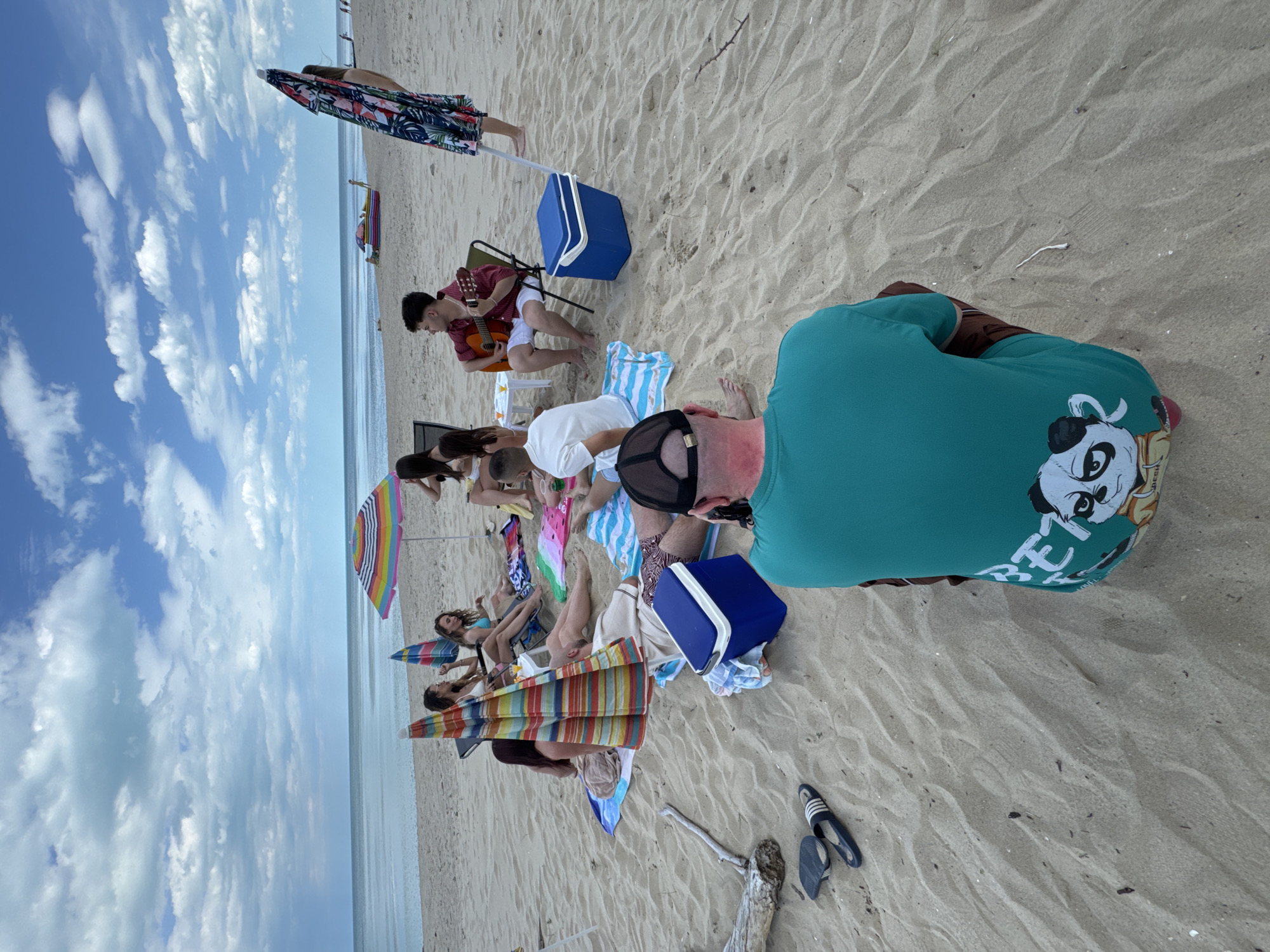 The image depicts a group of people relaxing on a beach. The location is likely Bliznatsi, Bulgaria, given the prompt context. The scene captures a sunny day with a visible sky and ocean in the background. Several individuals are visible. One man is playing a guitar. Some people are sitting under umbrellas and appear to be sunbathing or relaxing. A person wearing a green shirt with a cartoon design is positioned closer to the camera. There are also beach accessories visible, such as coolers, beach towels, and flip-flops scattered around. The overall impression is of a relaxed and casual beach scene.