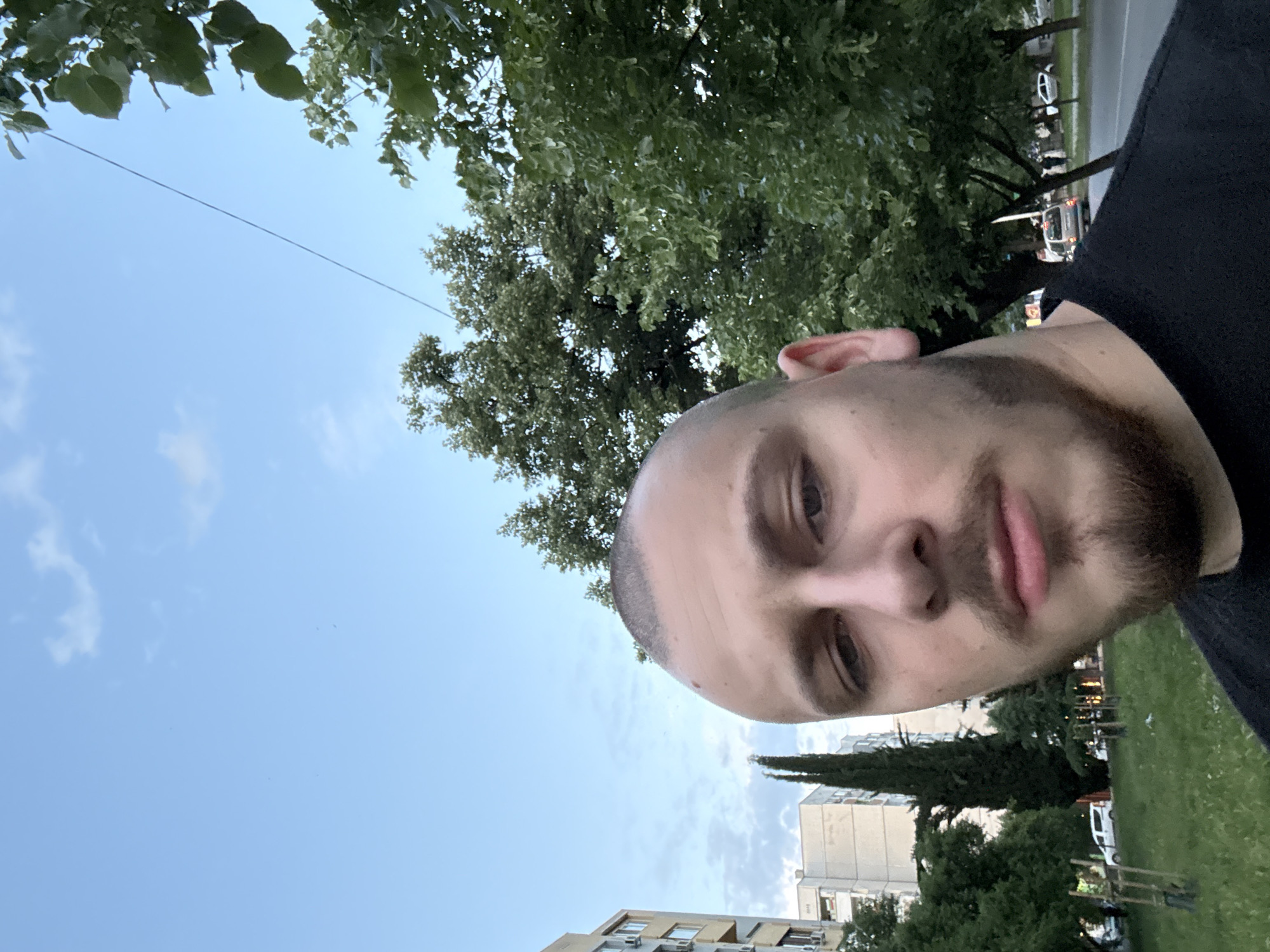 A close-up photograph shows a man's face, with the background showing an outdoor scene in Stara Zagora, Bulgaria. The man has a close-cropped haircut and a beard. In the background are trees, buildings, and a blue sky with clouds. There is also a road with a vehicle. The photo is taken from a low angle.