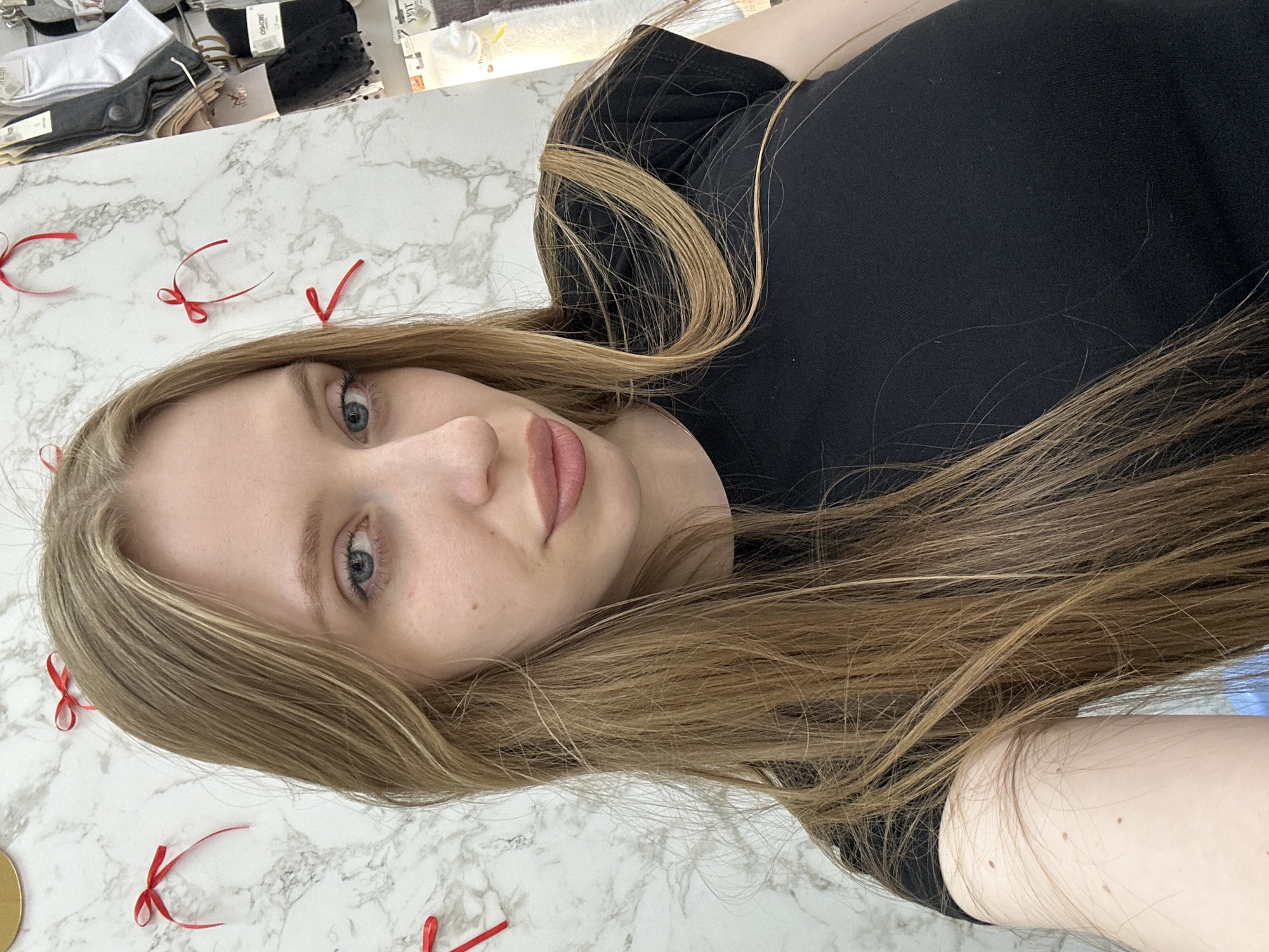 A woman with long, light brown hair and fair skin is lying on a marble-patterned surface. She is wearing a black top. Red ribbon bows are scattered across the surface. Behind her head are rows of packaged socks or other clothing items. The location, Chaikovsky, Russia, cannot be determined from the image.
