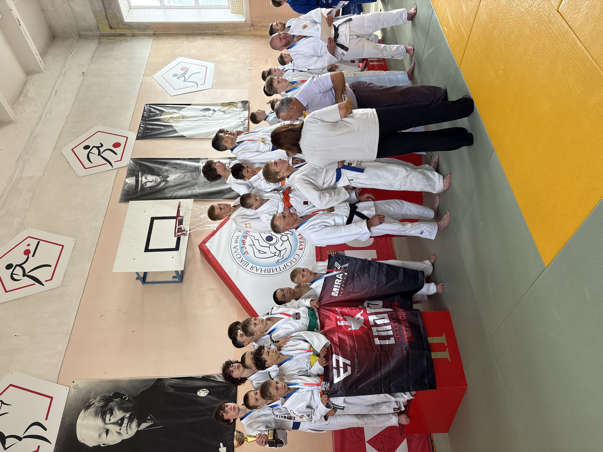 A group of approximately sixteen young male athletes and five adults are gathered for an awards ceremony in a gymnasium located in the Sports School named after V. R. Kuyeda, Russia, specializing in Judo. The floor is covered with green and yellow tatami mats.
On the left, a red two-tiered podium, marked with a gold Roman numeral 'II' on the lower tier, supports approximately twelve young males. These athletes, ranging from pre-teen to late teens, wear white judogi with various colored belts including white, yellow, green, and blue. Most have medals, predominantly gold- and silver-colored, around their necks. The leftmost athlete on the highest tier holds a gold-colored trophy. In the foreground, six of these athletes hold a large black banner with white and red lettering, featuring "EZ GROUP," a "Judo" logo depicting a throw, "MIRA SAFETY," and "СПОРТ ЗАПАДНЫЙ МБУ ДО СШ" (SPORT WESTERN MBOU DO SS).
To the right of the podium, an awards presentation is underway. A woman in a white top and black pants faces a man in a light purple collared shirt and dark pants, who appears to be presenting an award or medal to a young athlete in a blue-belt judogi. Further to the right, two adult men in judogi stand on the floor; one wears a black belt and holds a white certificate or paper, while the other is in a blue judogi. An additional adult man in a black-belt judogi is partially visible on the far left, behind the podium.
The background features light peach-colored walls with exposed white ceiling beams. Three large black and white portrait banners hang on the wall: Jigoro Kano (founder of Judo) on the left, a man in a fedora in the center, and another martial artist in a gi on the right. Four hexagonal plaques depicting stylized sports figures (table tennis, soccer, and two grappling sports) are mounted above these portraits. A white basketball backboard and red hoop are centrally positioned on the wall behind the ceremony. A large triangular banner, predominantly red and white, displaying "СПОРТИВНАЯ ШКОЛА ИМ. В. Р. КУЕДА ДЗЮДО" (Sports School named after V. R. Kuyeda Judo) and a stylized fish/dragon logo, is mounted to the right of the basketball hoop. Natural light enters through a large window on the far right, covered by a metal mesh screen.