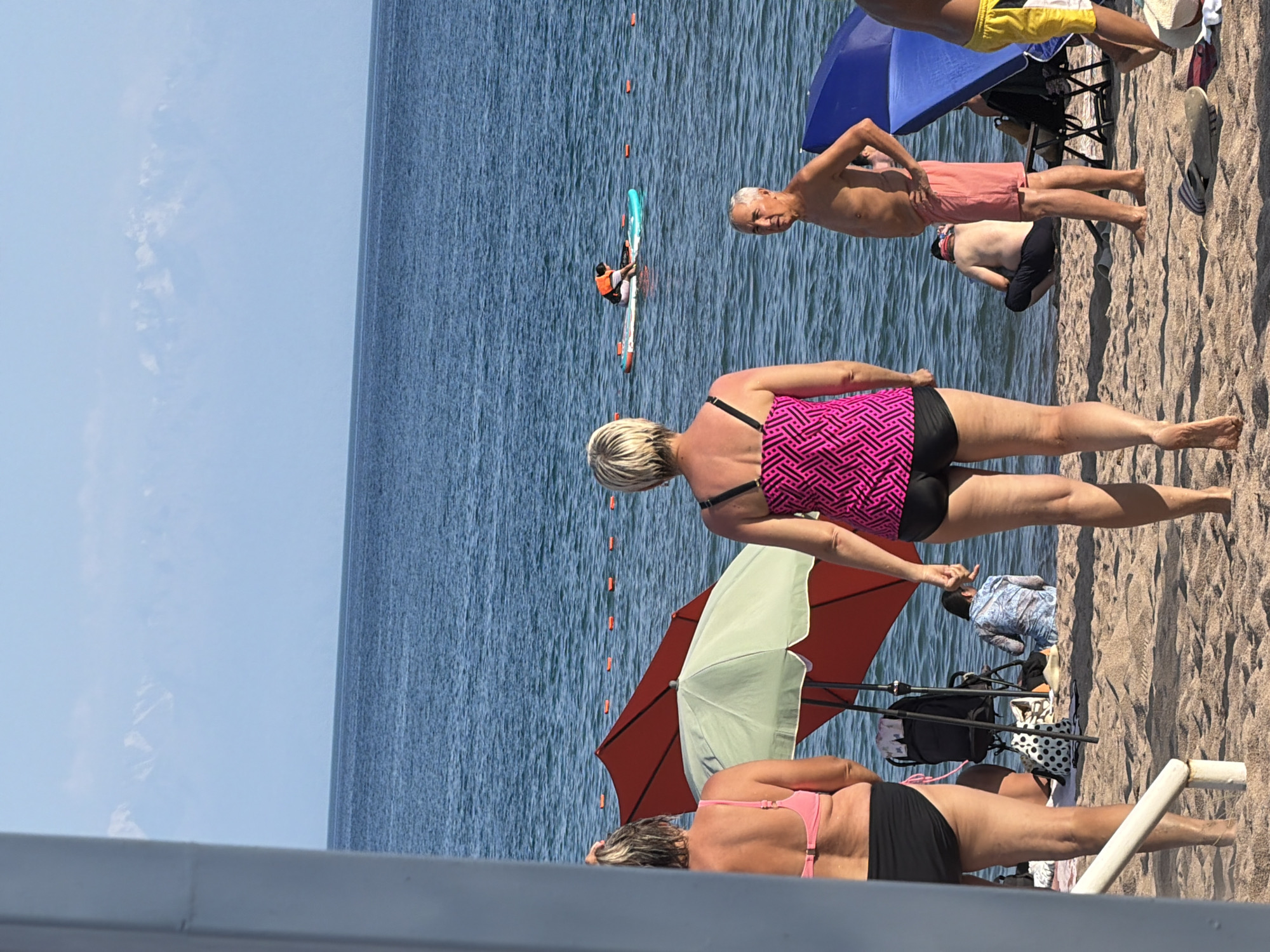 The image depicts a beach scene with several individuals. In the foreground, two women are visible from behind, wearing swimwear. One woman in a pink top and black bottoms, and the other in a pink patterned tankini and black bottoms. Further back, an older man with grey hair is standing, wearing pink shorts. A person in the water is on a paddleboard, wearing an orange life vest. There are beach umbrellas, chairs, and beach bags on the sand. The background shows a large body of water with distant mountains under a clear blue sky. The overall scene suggests a leisurely day at the beach.