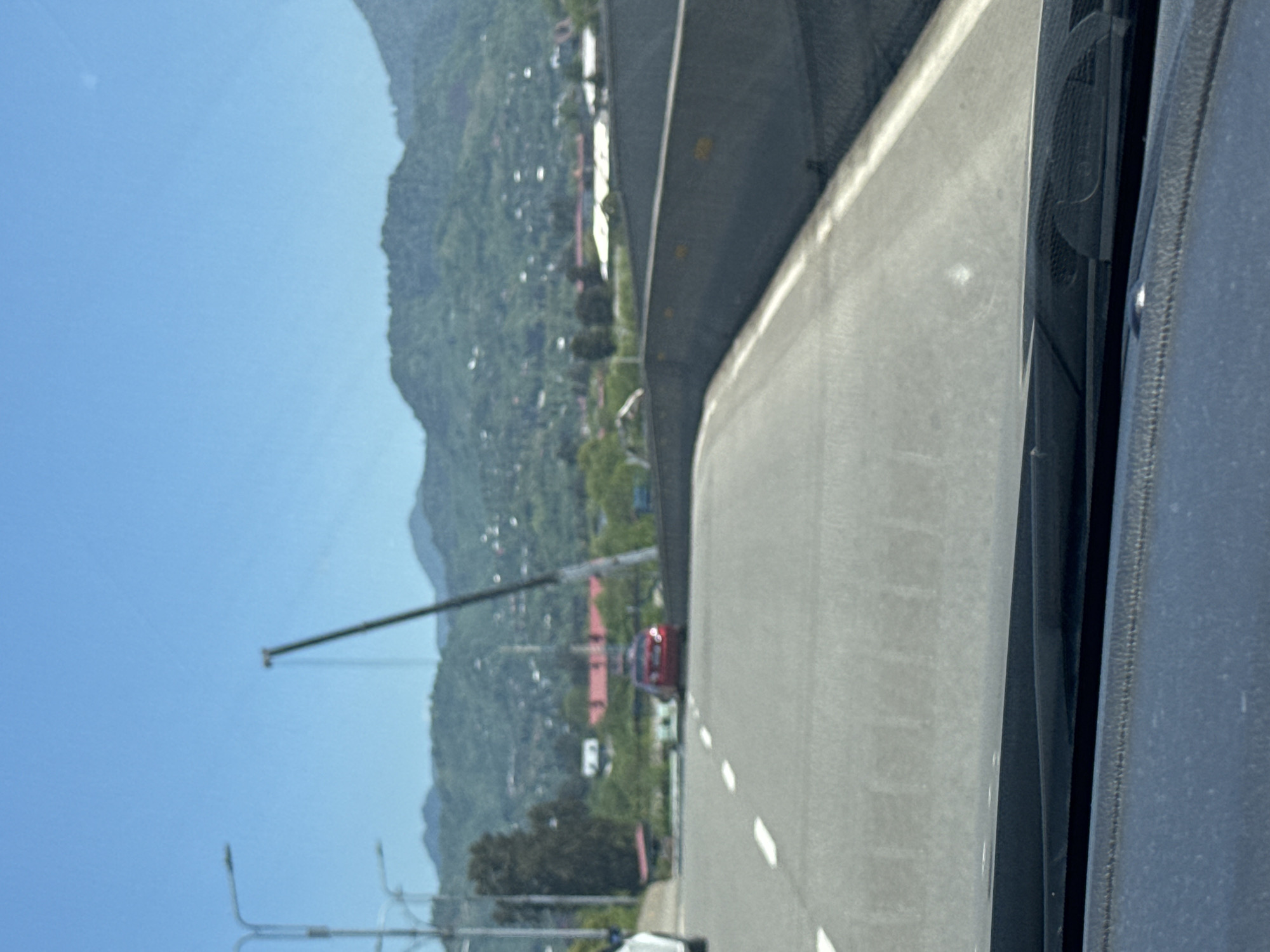 The image, taken from inside a vehicle, shows an elevated road curving to the right. The road is gray and has white lane markings. To the left of the road is a view of a mountain covered in green vegetation, with buildings scattered across the landscape. In the distance, a tall construction crane is visible against a clear blue sky. The interior of the vehicle is visible on the right side of the frame, with parts of the dashboard and window frame appearing in dark gray. The scene is likely located on the outskirts of the city of Batumi, Georgia, based on the hilly terrain and the combination of urban and natural elements.
