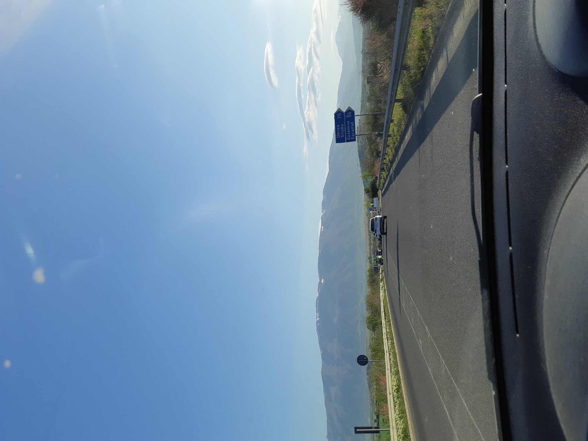 The image captures a bright, sunny daytime view from inside a vehicle traveling on a highway in Bulgaria, consistent with the Marino Pole location. On the right, a dark portion of the car's interior, possibly the dashboard or A-pillar, is visible. The asphalt road stretches into the distance, marked with white lines, with a metal guardrail along its right side.
Beyond the guardrail, green vegetation lines the road. In the mid-ground, a blue road sign provides directions and distances in Bulgarian and English: "Шипка Shipka 70" and "Казанлък Kazanlak 55". Several other vehicles are visible on the road ahead.
The background features a grand mountain range with some snow-dusted peaks, extending across the horizon under a clear, vibrant blue sky dotted with a few elongated white clouds. The scene conveys a sense of a pleasant road trip through a scenic, mountainous landscape under excellent weather conditions.
