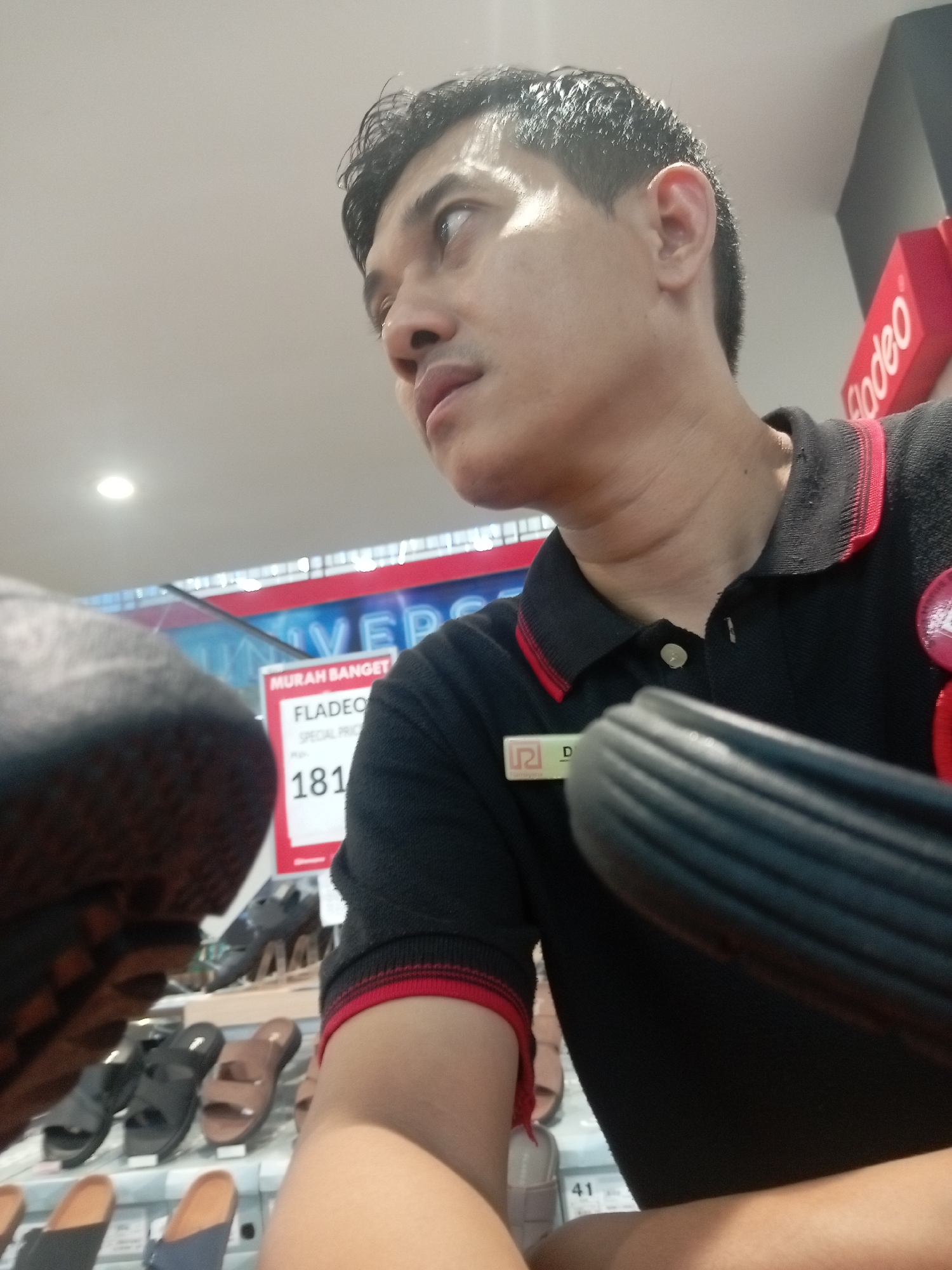 A man is captured in a low-angle, medium shot, looking upwards and to the right. He is wearing a black polo shirt with red trim on the collar and sleeves. A name tag is visible on the left side of his shirt. The man's right arm is in the foreground, obscuring part of the scene. Behind him, shelves are filled with various footwear, including sandals. A red sign with white text, including "MURAH BANGET" (very cheap) and "SPECIAL PRICE" with the number "181", is prominently displayed. In the background, a sign with the word "UNIVERSE" is visible, along with elements of a retail environment, including what appears to be an escalator. The overall setting suggests a shoe store in Batam, Indonesia.