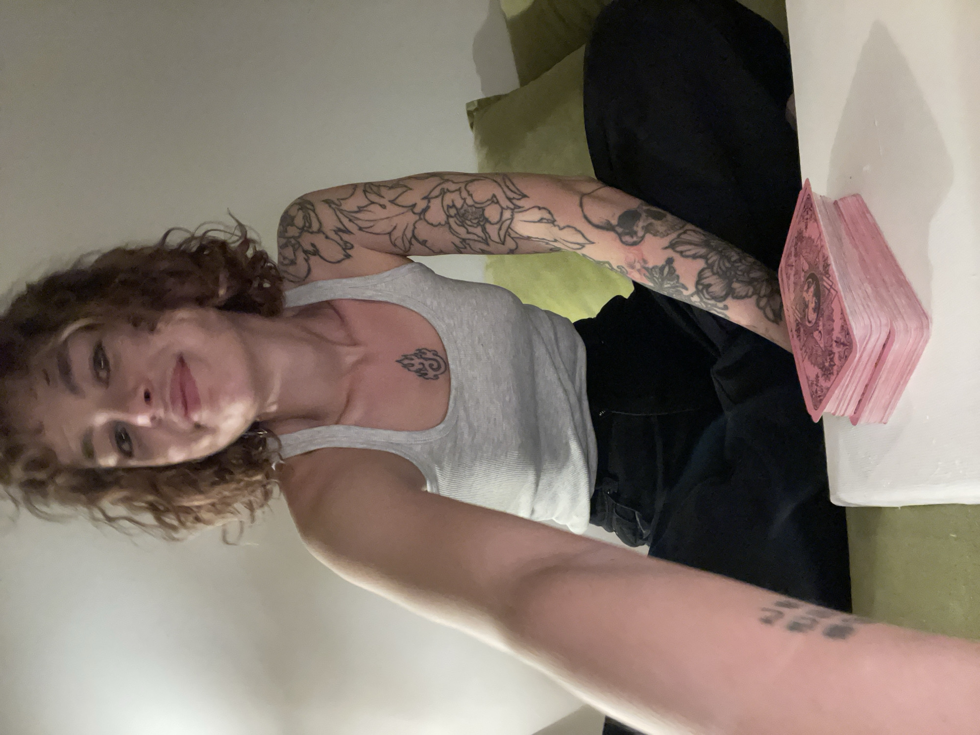 A woman with curly brown hair, a gray tank top, and black pants is on an olive green couch with a stack of pink tarot cards on a white surface next to her. She has tattoos on both arms and her chest. The location is not discernible as Boreti, Montenegro.