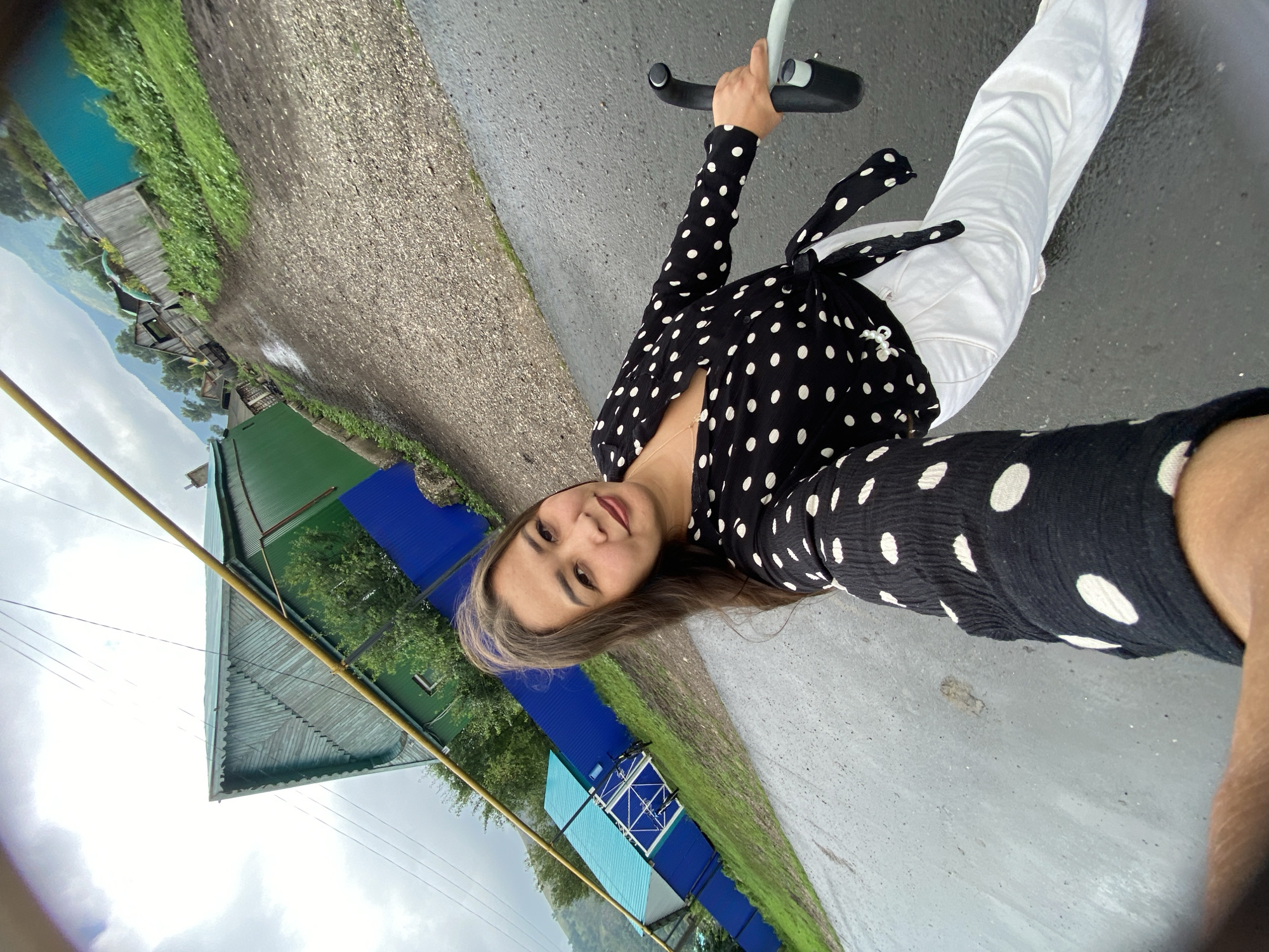 A woman is taking a selfie outdoors in Maloarslangulovo, Russia. She is standing on a wet gravel and asphalt road. She is wearing a black and white polka dot blouse with a tie at the waist and white pants. In her right hand, she is holding onto the handlebars of a bicycle or stroller. Behind her, there are several houses with green and blue fences, some trees, and a mountain in the distance. The sky is overcast.