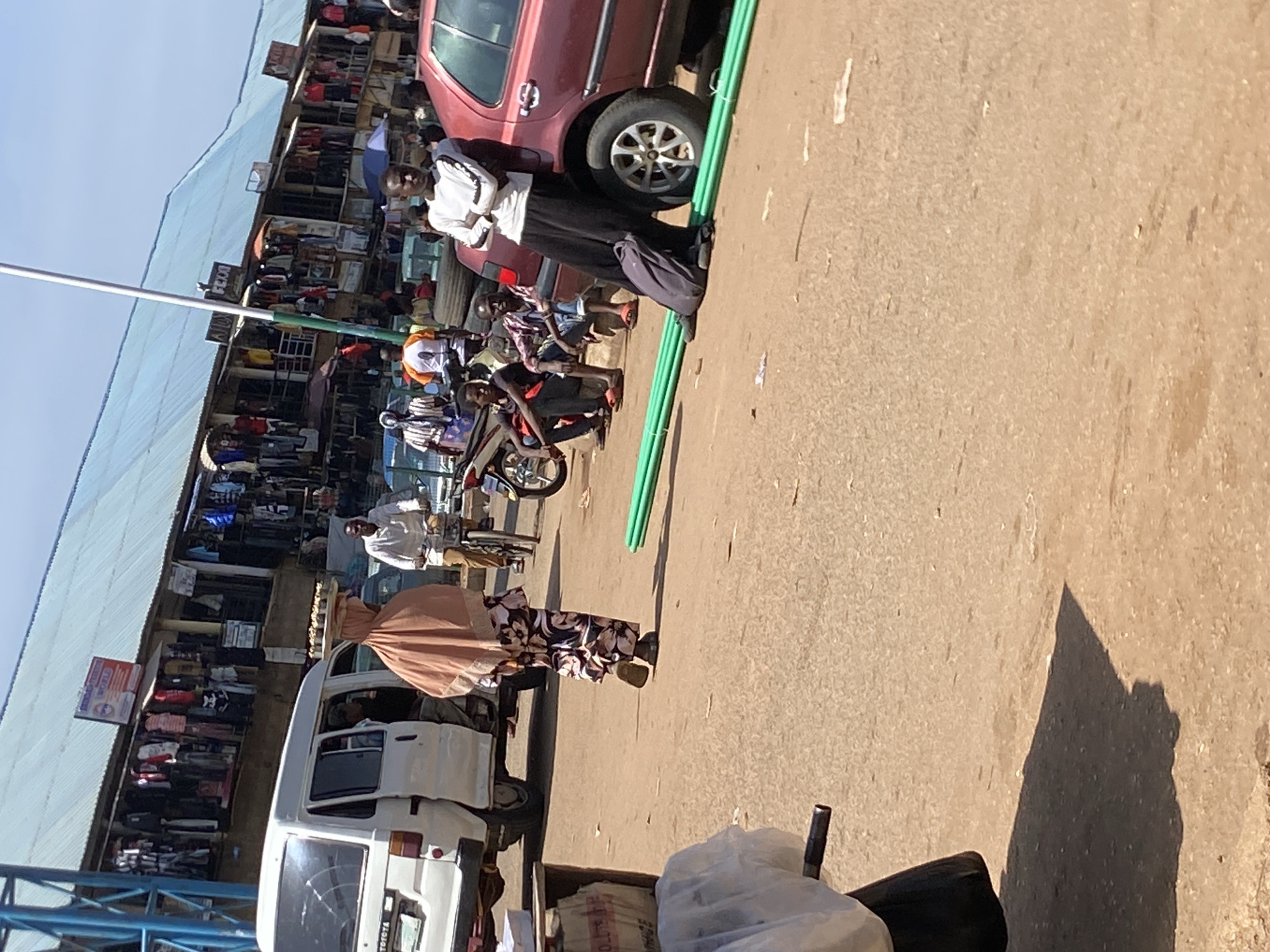 The image depicts a bustling marketplace scene in Bukuru, Nigeria, under a bright, clear sky. A white van is parked on the left, its side door ajar. To its right, a woman wearing a light brown headscarf and a patterned dress walks away from the camera, carrying a flat tray of what appears to be food or goods on her head. In the background, a row of open-fronted shops lines the street, displaying a variety of clothing and merchandise. Several people are visible in and around the shops, including a man on a bicycle in the middle ground. Further to the right, a group of men are seated and standing near a red car. One man in a white long-sleeved shirt is leaning against the car with his arms crossed, and a dark backpack rests at his feet. In the foreground, a collection of green cylindrical pipes lies on the dusty ground. The overall environment suggests a busy commercial hub with a mix of transportation, retail, and pedestrian activity.
