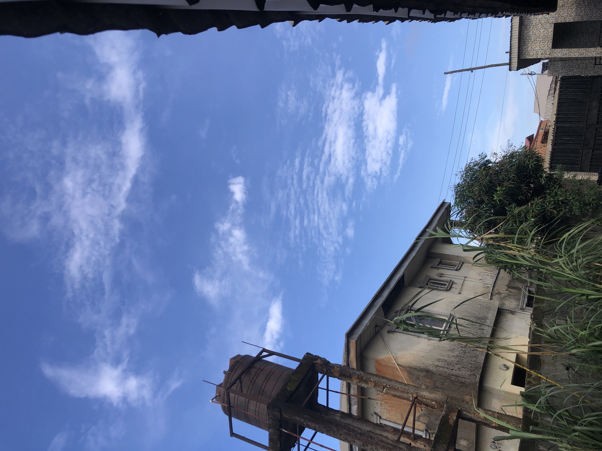 In Ado, Nigeria, the image depicts an outdoor scene with a bright blue sky filled with scattered, wispy white clouds. In the foreground, tall, green grasses grow along a concrete wall. To the left, an old, weathered building with a damaged water tank on a rusty metal and concrete support stands prominently. The building's facade is stained and appears to be in disrepair, with visible windows and piping. Adjacent to this, a section of a roof with dark tiles is visible on the right side of the frame, suggesting another structure. Further in the background, glimpses of other buildings with corrugated roofs and a dark metal gate topped with barbed wire are visible. Power lines stretch across the sky above some of the structures. The overall impression is one of an urban environment with elements of aged infrastructure.