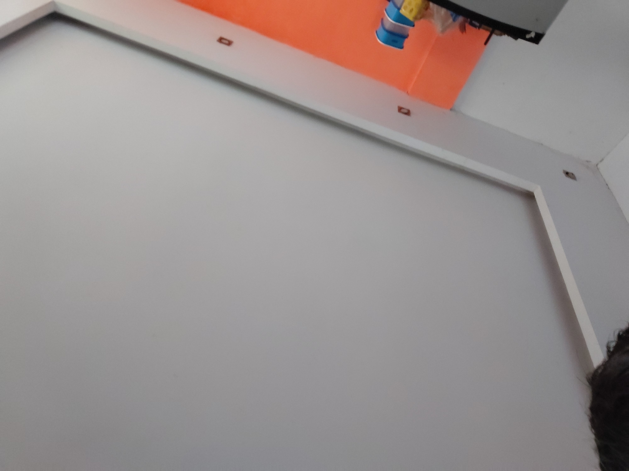 The image captures an upward-angled view of an indoor space, likely a room, in Santa Ana, Venezuela. A significant portion of the frame is dominated by a plain light grey wall. Running horizontally across the upper part of this grey wall is a white architectural trim or frame, which appears to be either a doorframe header, a window frame, or decorative wall molding.
Above this white trim, an orange-colored surface is visible, possibly another wall or a large structural element. Several small, dark, rectangular features are sparsely distributed on both the white trim and the orange surface; these could be electrical outlets, mounting brackets, or embedded fixtures.
In the upper-left corner, a cluster of indistinct colorful objects, including blue and yellow items, is partially visible, seemingly hanging from something out of frame. Towards the top-right, a dark, rectangular object, possibly a monitor, TV, or shelf, is partially visible, with smaller items hanging from its underside. A small section of dark, curly hair is visible in the bottom-right corner, suggesting the photographer's head is partially in the shot. The lighting indicates an indoor setting, with no specific time of day or external weather conditions discernible. There is no visible text in the image.
