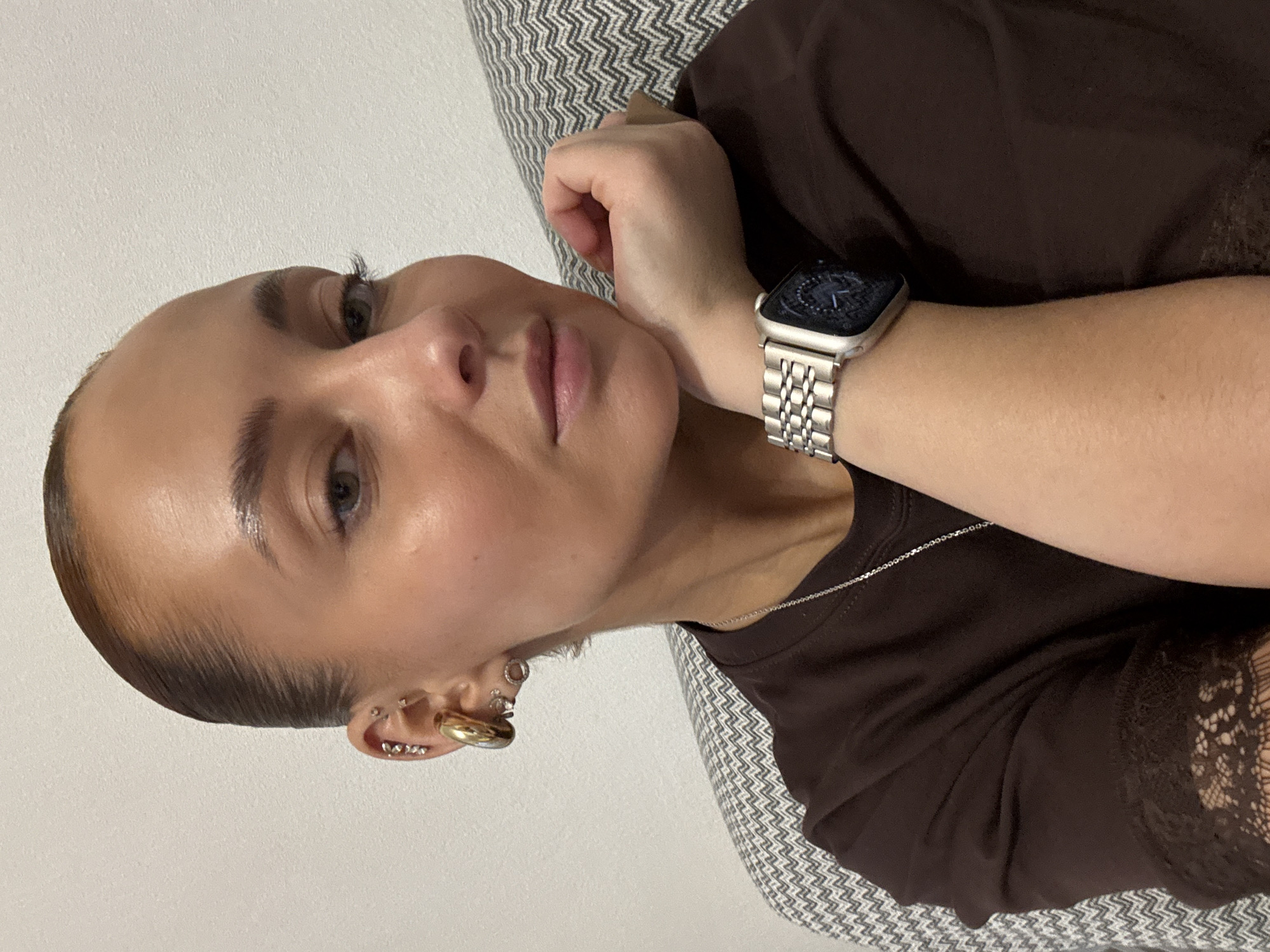 A woman with fair skin, short, slicked-back brown hair, and multiple ear piercings is seen in a close-up, slightly angled view. She is wearing a dark brown t-shirt with lace detailing and a silver necklace. A silver Apple Watch is on her left wrist, resting on her forearm. The background includes a gray and white patterned cushion or pillow behind her. There is no indication of the location being in the city of Chaikovsky, Russia.